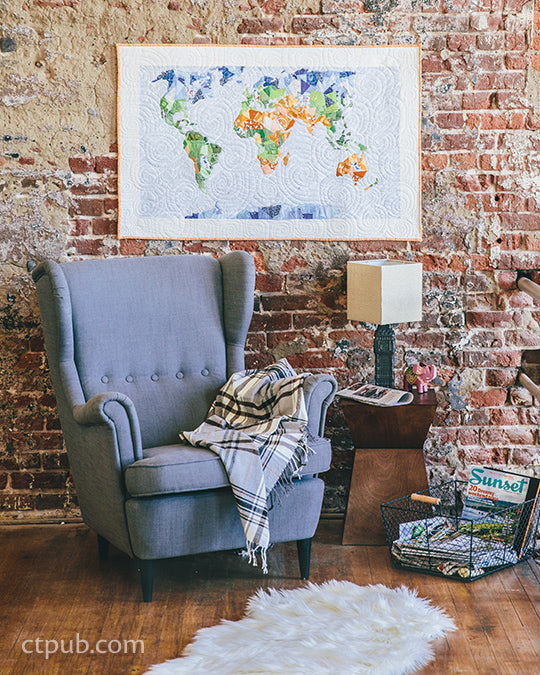 A cozy reading nook with a gray armchair, a plaid blanket, and a small wooden table with a lamp. On the brick wall hangs C&T Publishing’s Scrappy Bits Appliqué Print-on-Demand Edition, featuring a colorful quilted world map design.