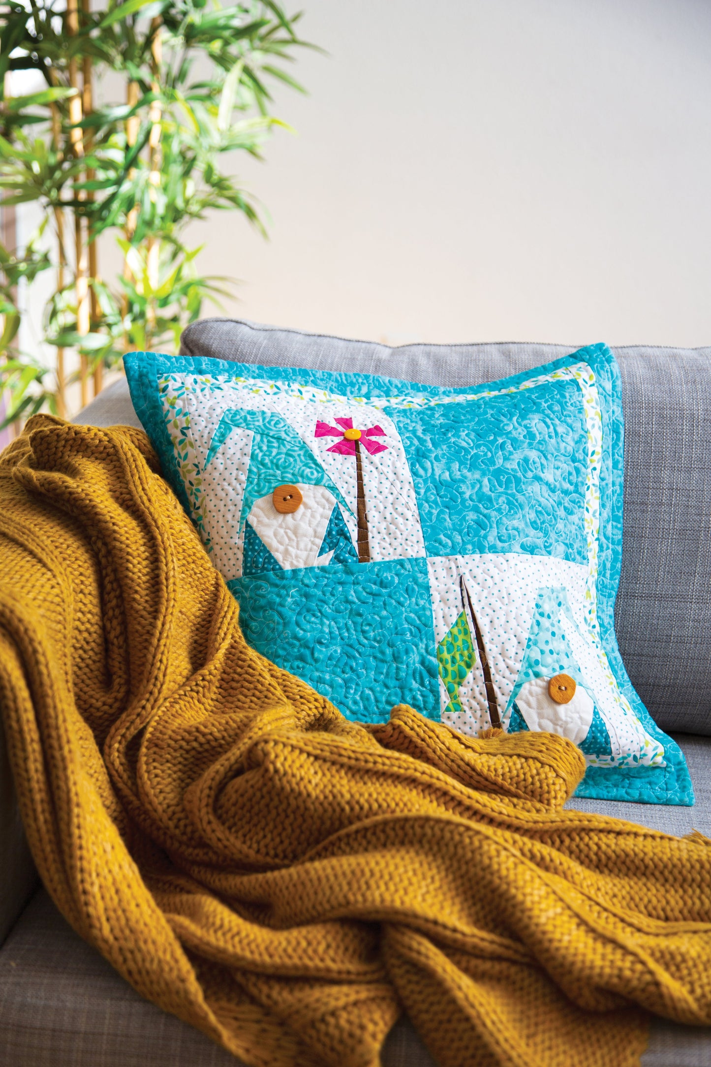 A gray sofa with a mustard yellow knitted blanket and a colorful pillow showing Mary Hertel's whimsical house and flower design from C&T Publishing's "Paper Piecing All Year Round" digital download. A green plant is in the background.
