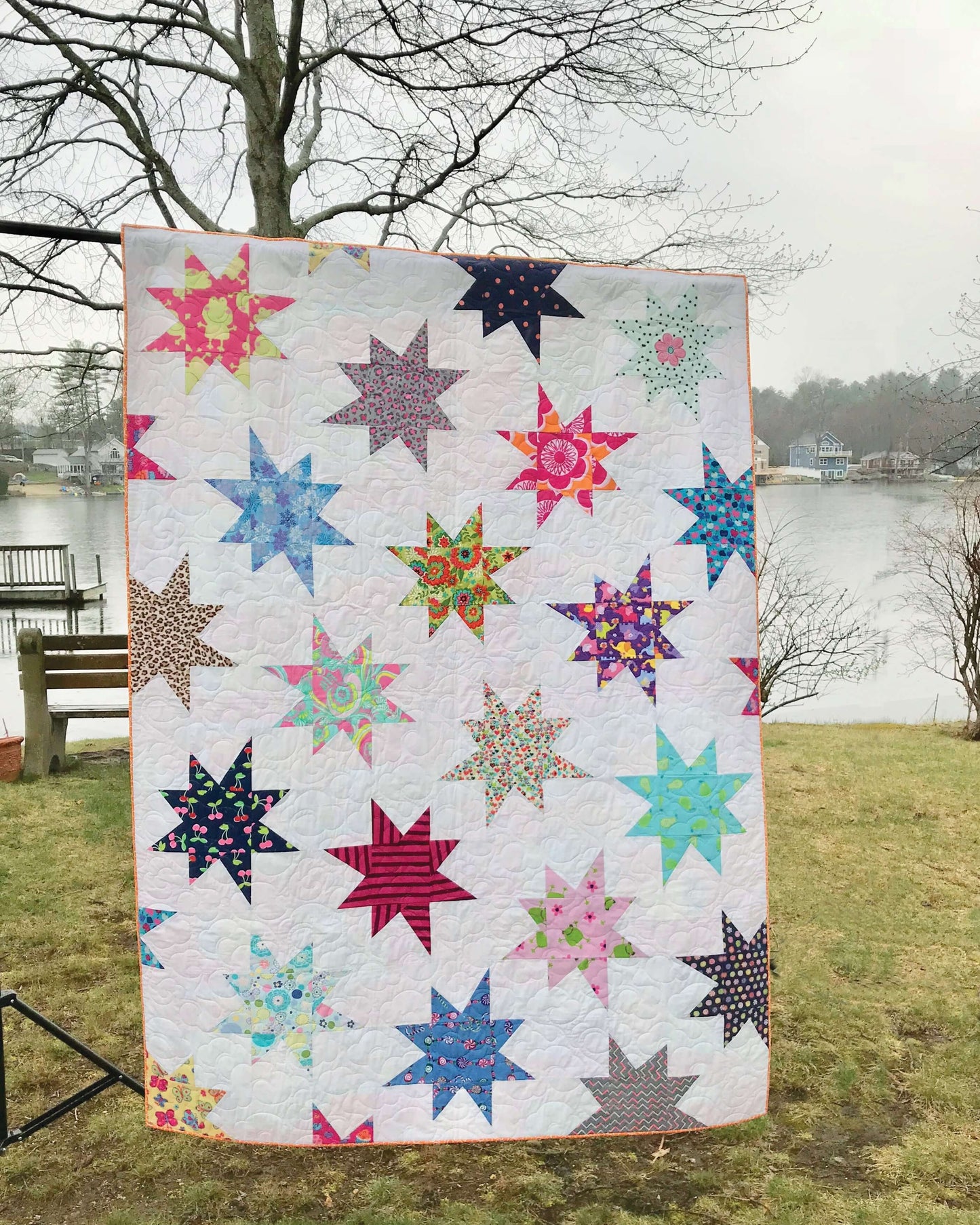 The Beyond the Tee—Innovative T-Shirt Quilts Digital Download by C&T Publishing is shown outdoors on a cloudy day, featuring multicolored star patterns with a scenic lake and bare trees in the background.