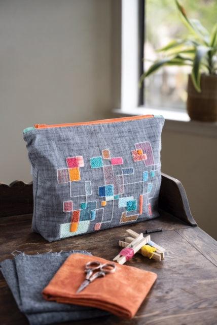 The Modern Crewel Embroidery Digital Download by Stash Books is shown as a gray pouch with colorful geometric embroidery, displayed on a wooden table beside fabric swatches, wool thread, scissors, and a potted plant by the window.