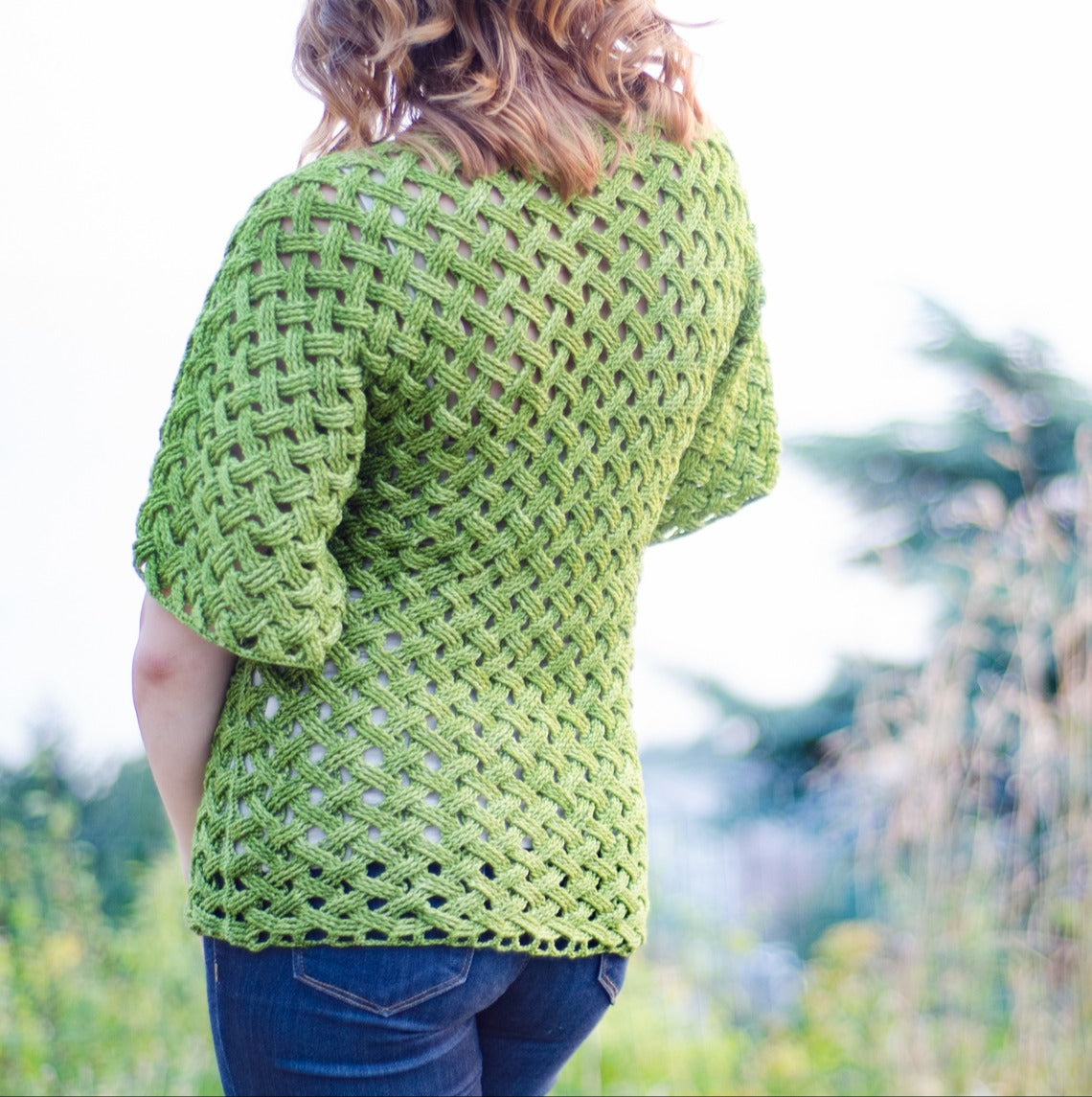 A person with wavy hair, facing away, wears a handmade Woven Cables Crochet top by Creative Spark. They stand outdoors amid greenery under a cloudy sky.