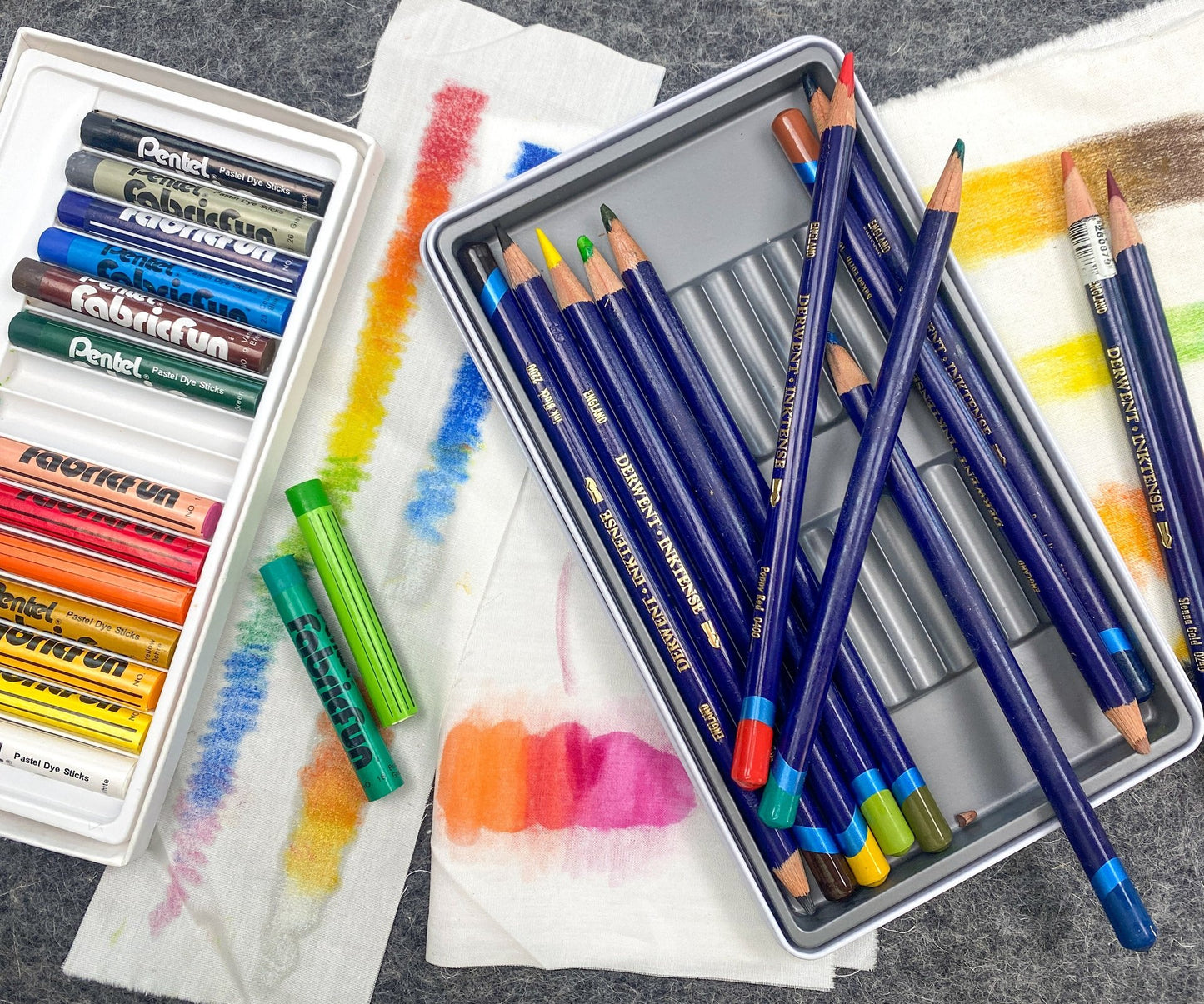 A set of Creative Sparks Coloring Fabric with Inktense Pencils and Pastels is displayed on the table. The pencils lie in a sleek metallic tin, and the pastels are in a white plastic case, with swatches on paper towels showcasing their potential for fabric coloring.