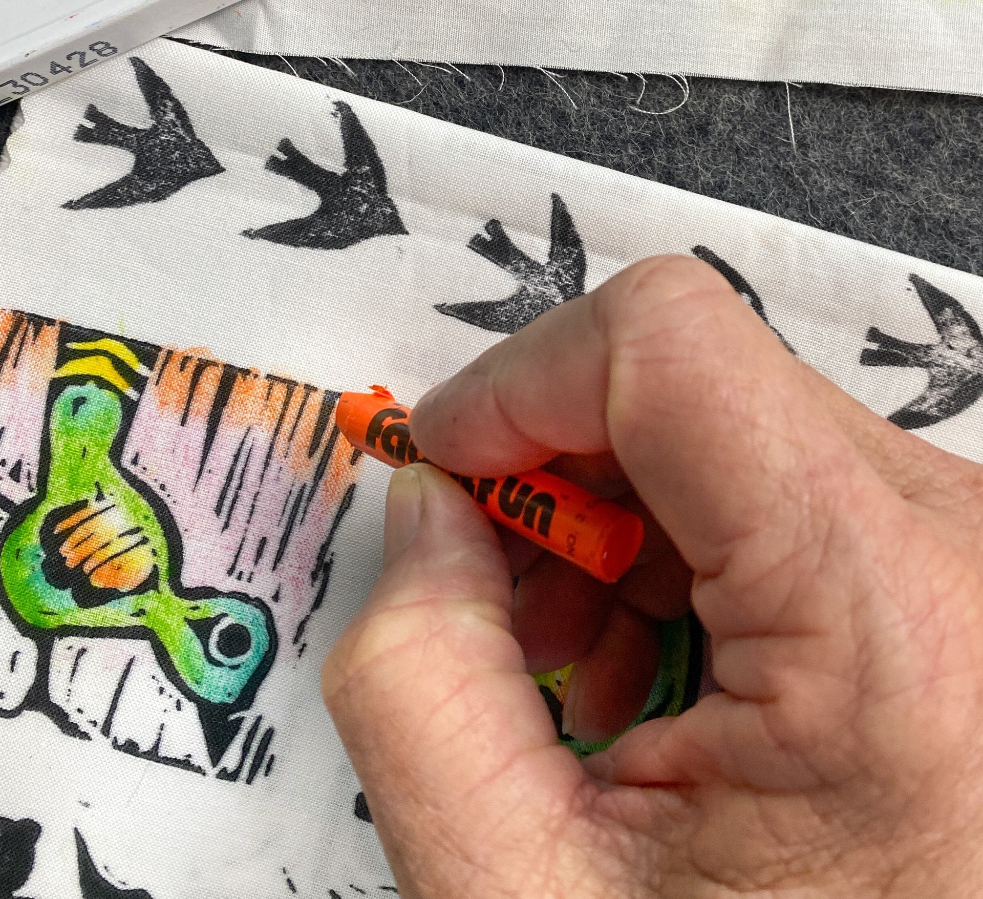 A persons hand applies Creative Sparks Inktense Pencils and Pastels to a fabric featuring black bird silhouettes and green patterns. The orange coloring highlights the artistry of fabric design, adding vibrant depth and texture.