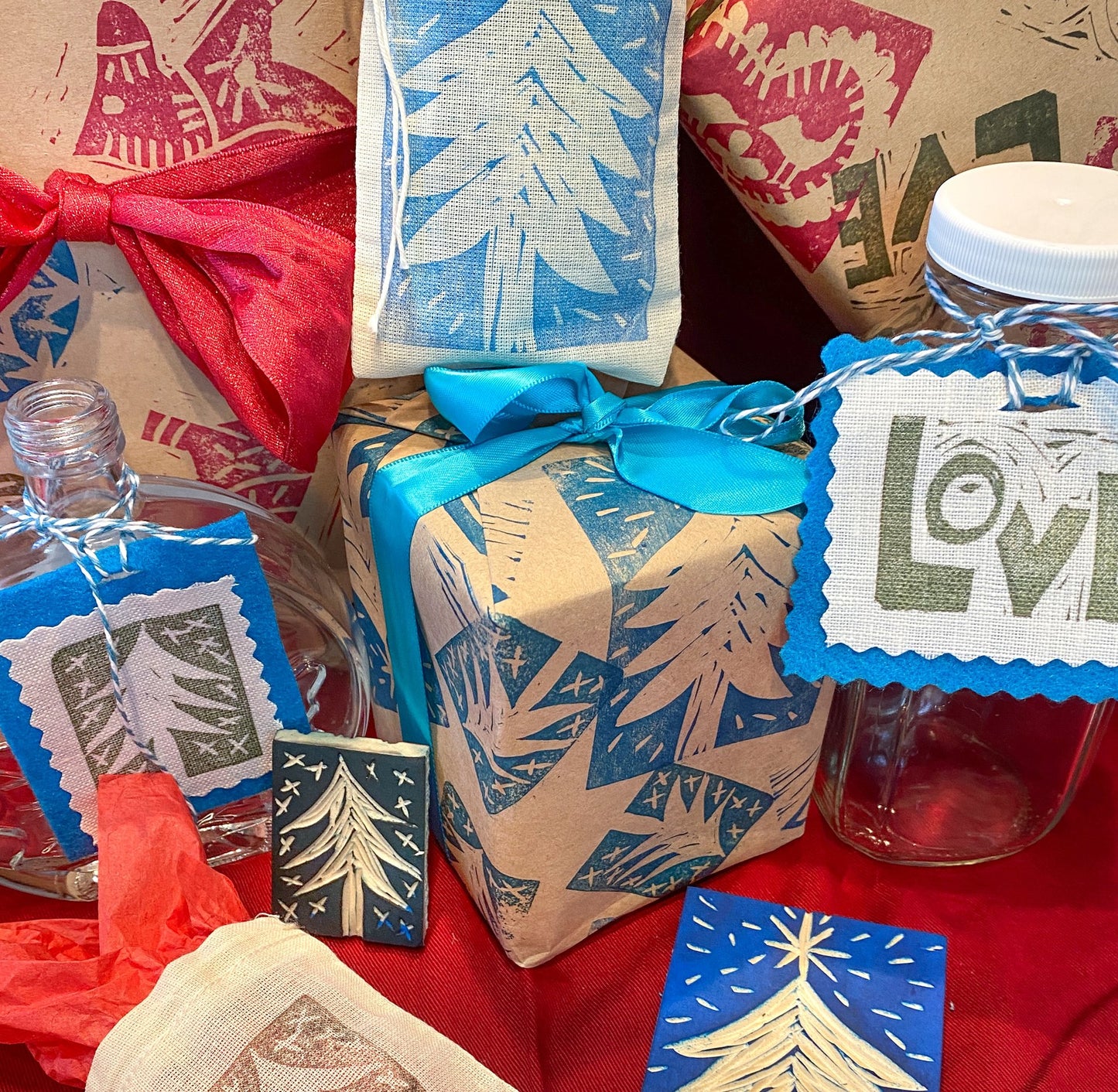 A festive display features "Block Printing: Design, Carve, Print" by Creative Spark, brown paper with blue and white tree prints, blue ribbons, glass bottles, and LOVE gift tags arranged on red holiday-themed fabric.