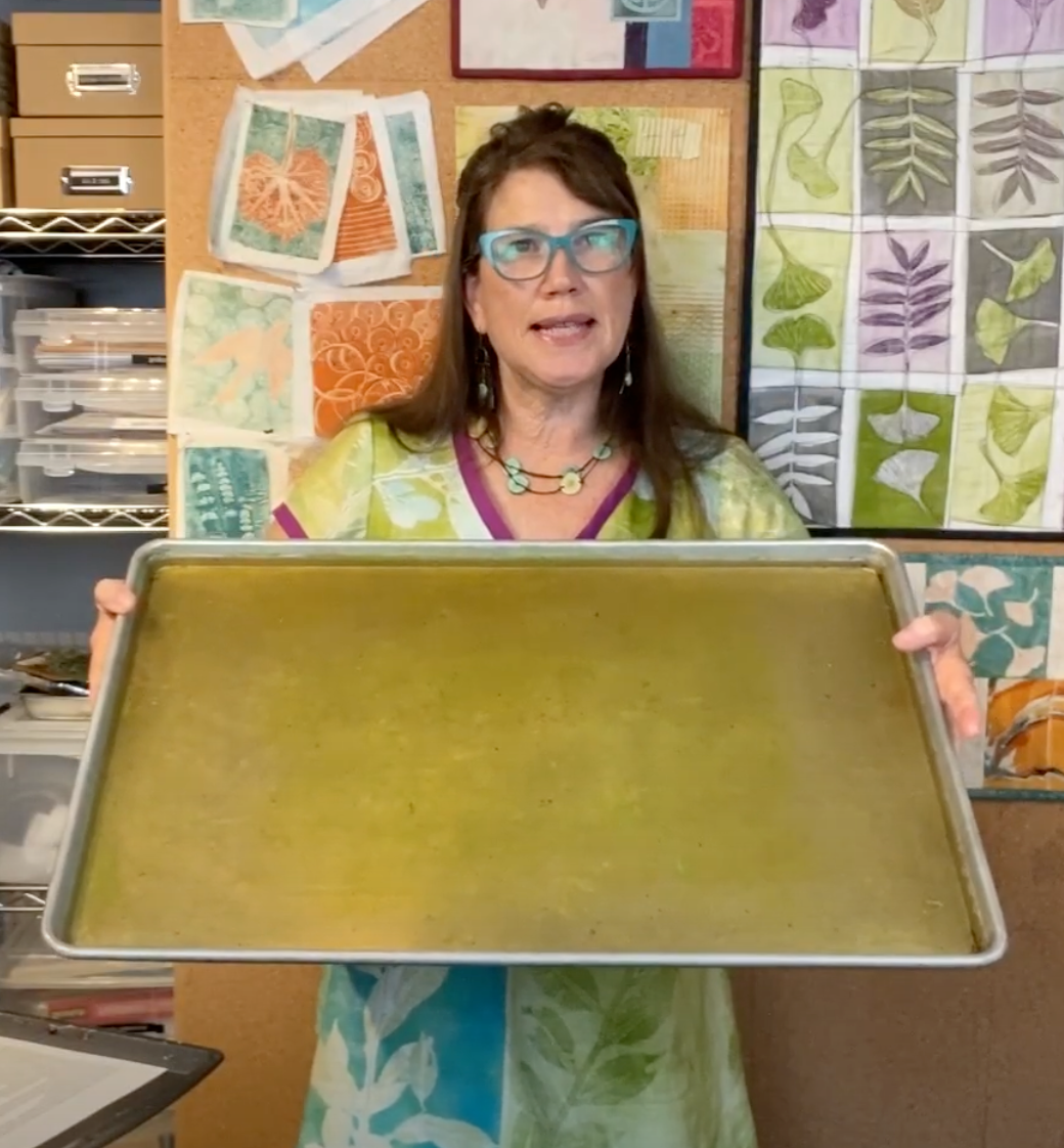 A woman in glasses and a colorful dress stands in an art studio, holding a large tray. Behind her are supplies and a wall decorated with botanical prints and textured samples made using Creative Spark’s Make Your Own Gel Printing Plate.