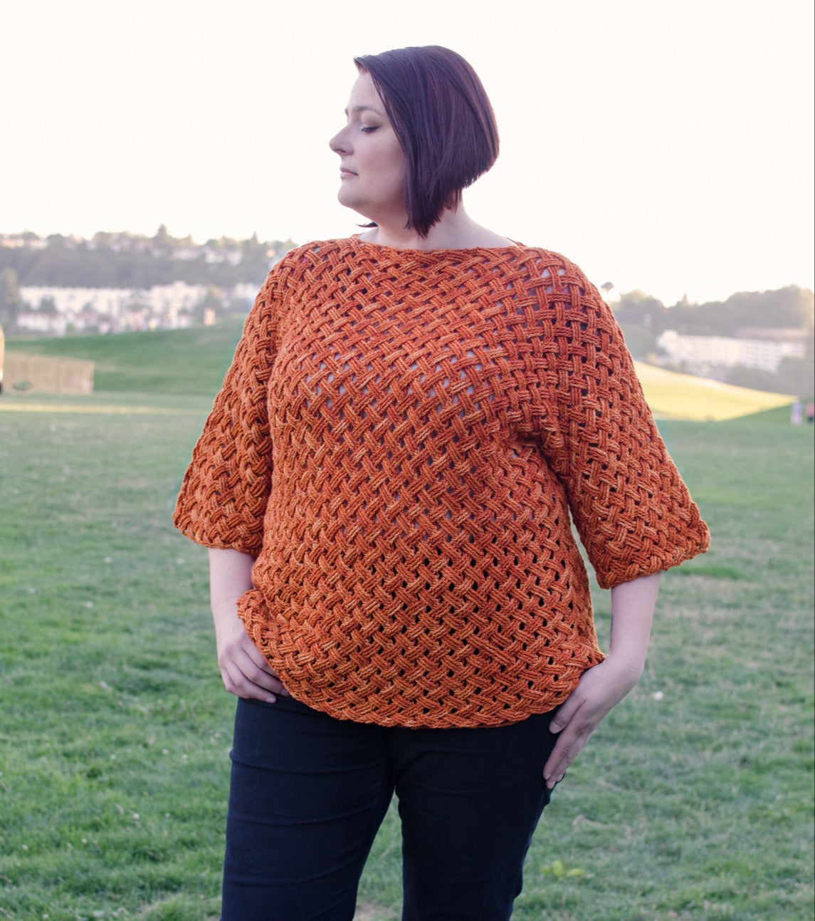 In a grassy field, a person with short dark hair wears an orange Woven Cables Crochet sweater by Creative Spark and black pants. They stand relaxed, gazing to the side, as trees and a clear sky offer a serene backdrop.