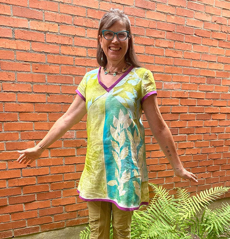 A smiling woman with glasses stands in front of a brick wall, wearing a leafy tunic she's made using the Creative Spark Make Your Own Gel Printing Plate, arms outstretched beside a green fern.