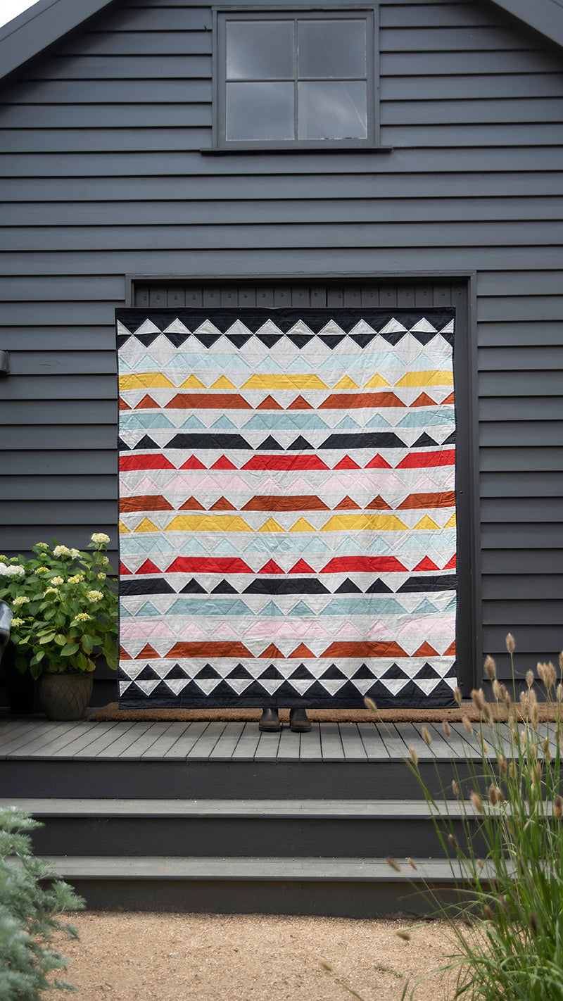 A vibrant geometric quilt from "Quilt Recipes" by C&T Publishing features rows of red, yellow, orange, black, and white triangles displayed on a dark gray wall with a potted plant on the left and steps leading to the door.