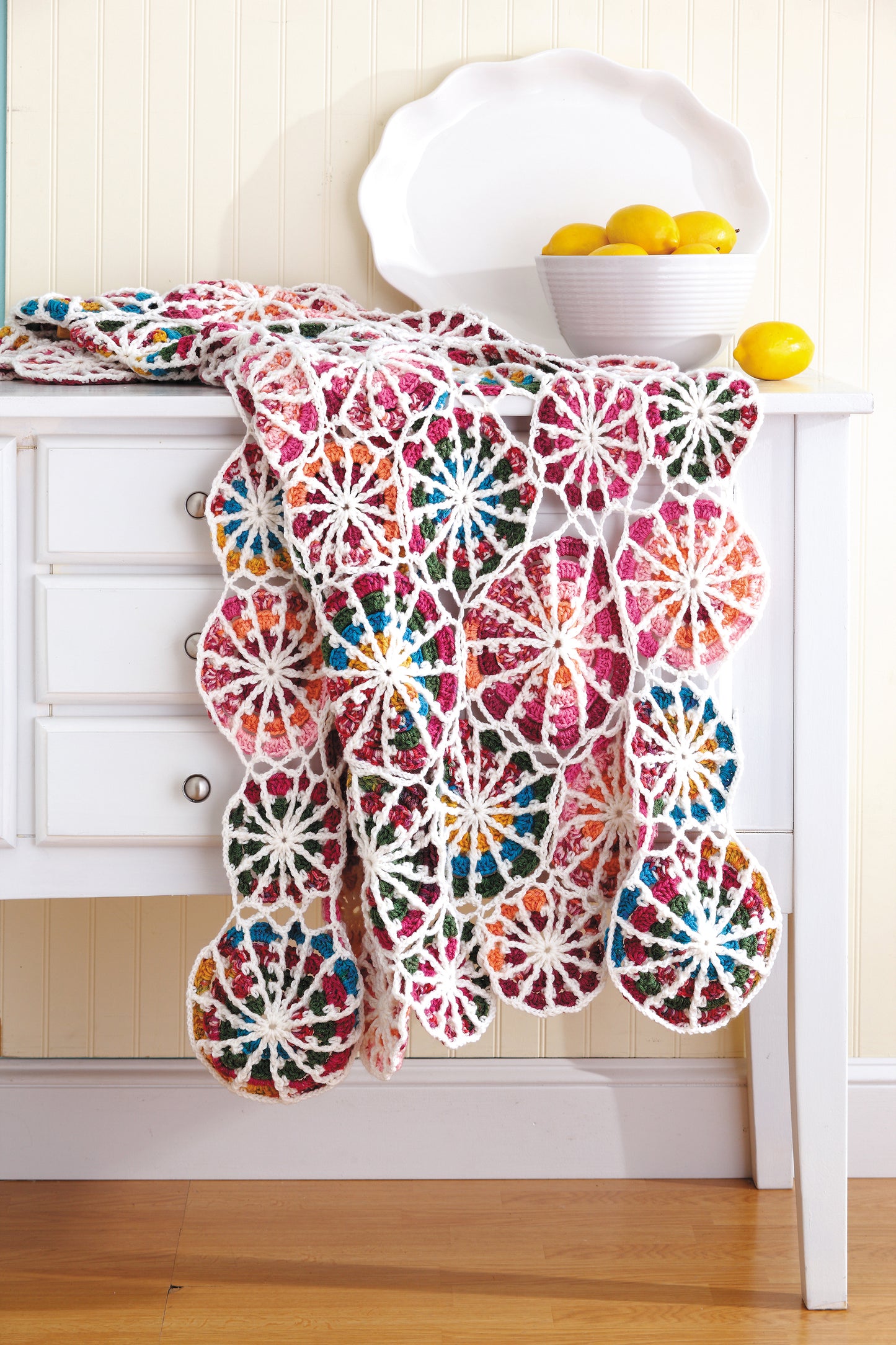 The "Everyday Crochet for All" blanket by Leisure Arts, featuring vibrant circular patterns ideal for beginners, is draped over a white table with a bowl of lemons, set against light wood paneling.