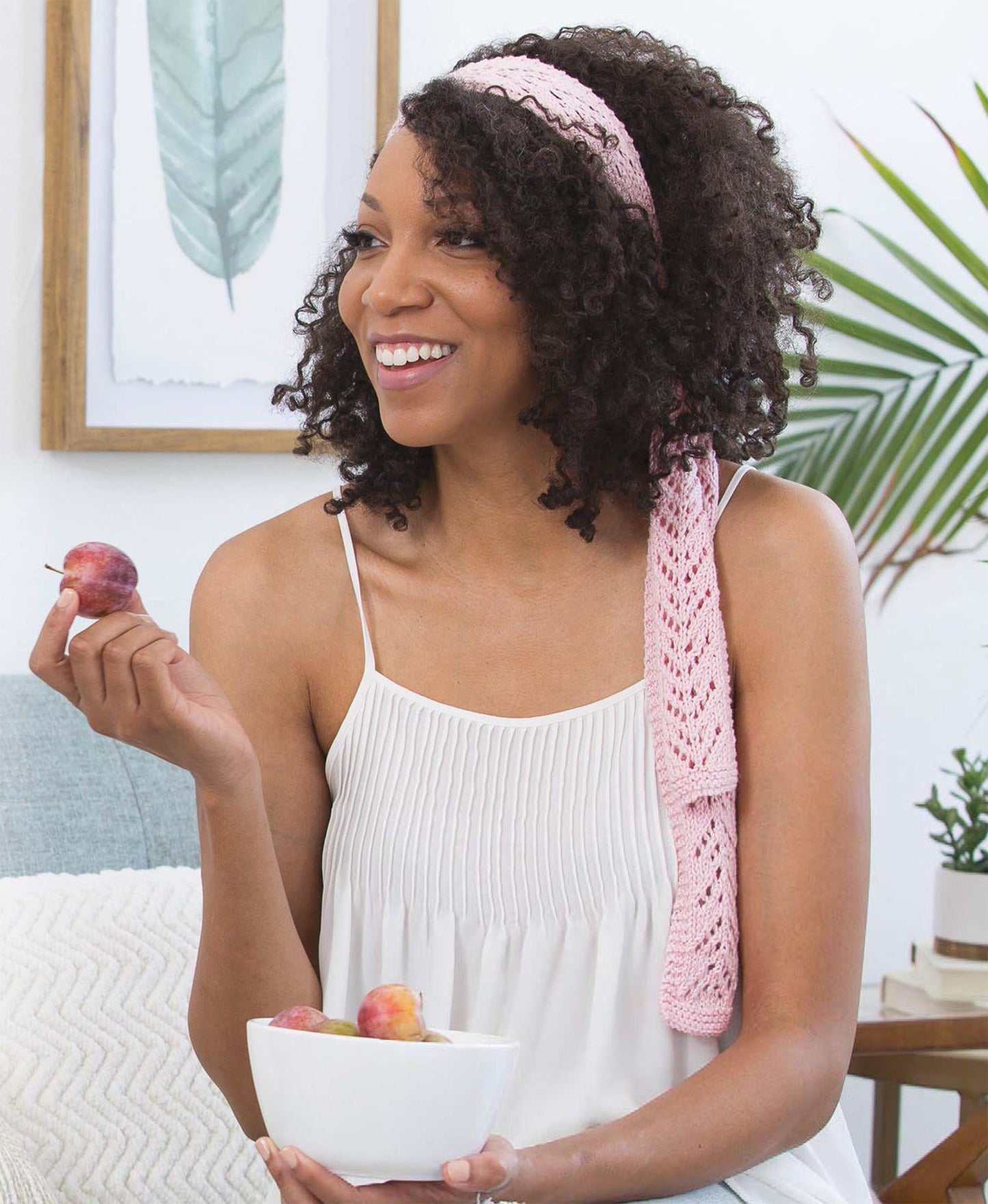 A smiling woman with curly hair and a pink headband relaxes on a couch, eating small plums and browsing "Get Your Lace On" by Leisure Arts—a collection of beginner women’s knitting patterns—amidst houseplants and framed art.