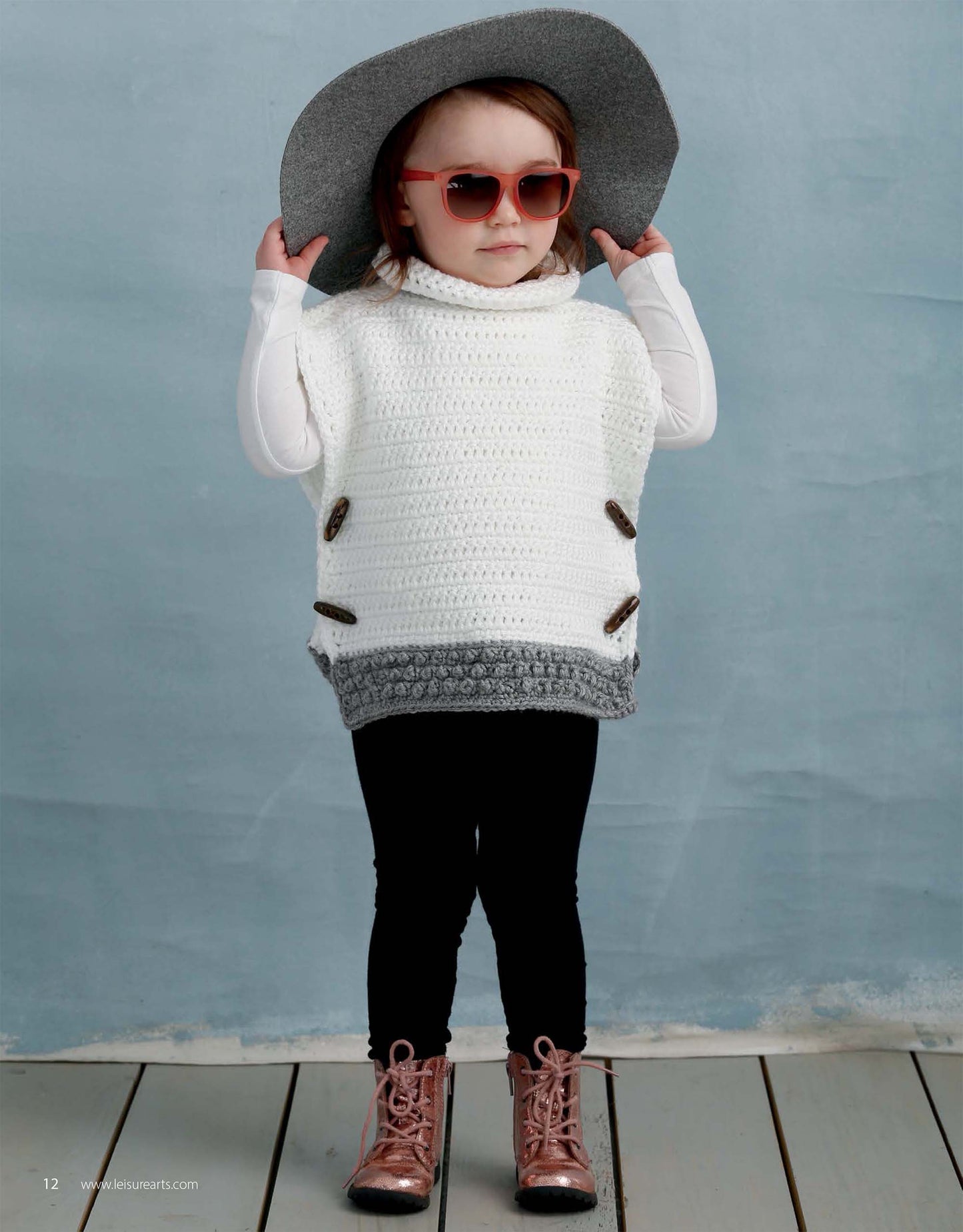 A young child in Leisure Arts' Crochet Lil' Hipsters knit sweater, large gray hat, pink sunglasses, black leggings, and shiny pink boots poses confidently against a blue background.