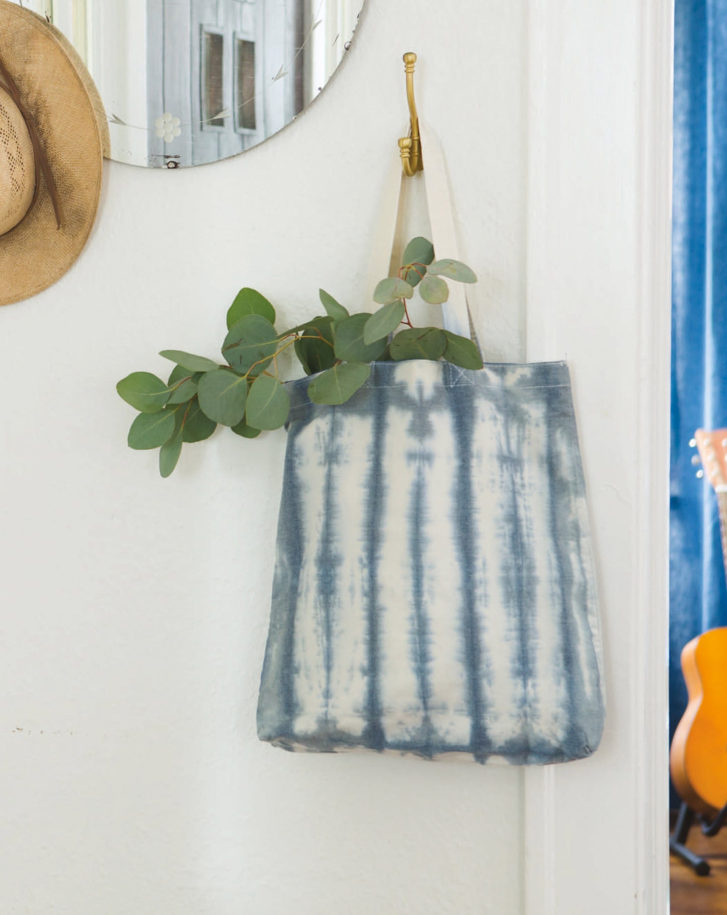 The "Shibori, Ice & Dip Dyeing" kit by Leisure Arts features a blue and white tie-dye tote bag for beginner dyeing on natural fibers. Hung with eucalyptus branches, it's styled with a mirror, straw hat, and an acoustic guitar nearby.