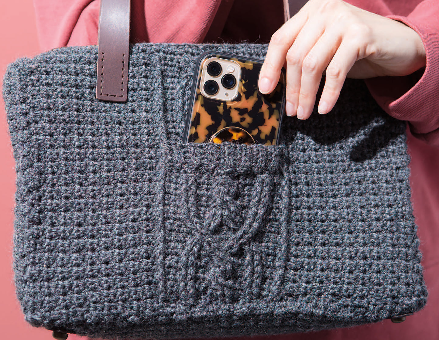 Someone slips a smartphone with a tortoiseshell-patterned case into the front pocket of a gray, hand-crocheted bag with brown handles from Leisure Arts’ Crochet Handcrafted Handbags collection.