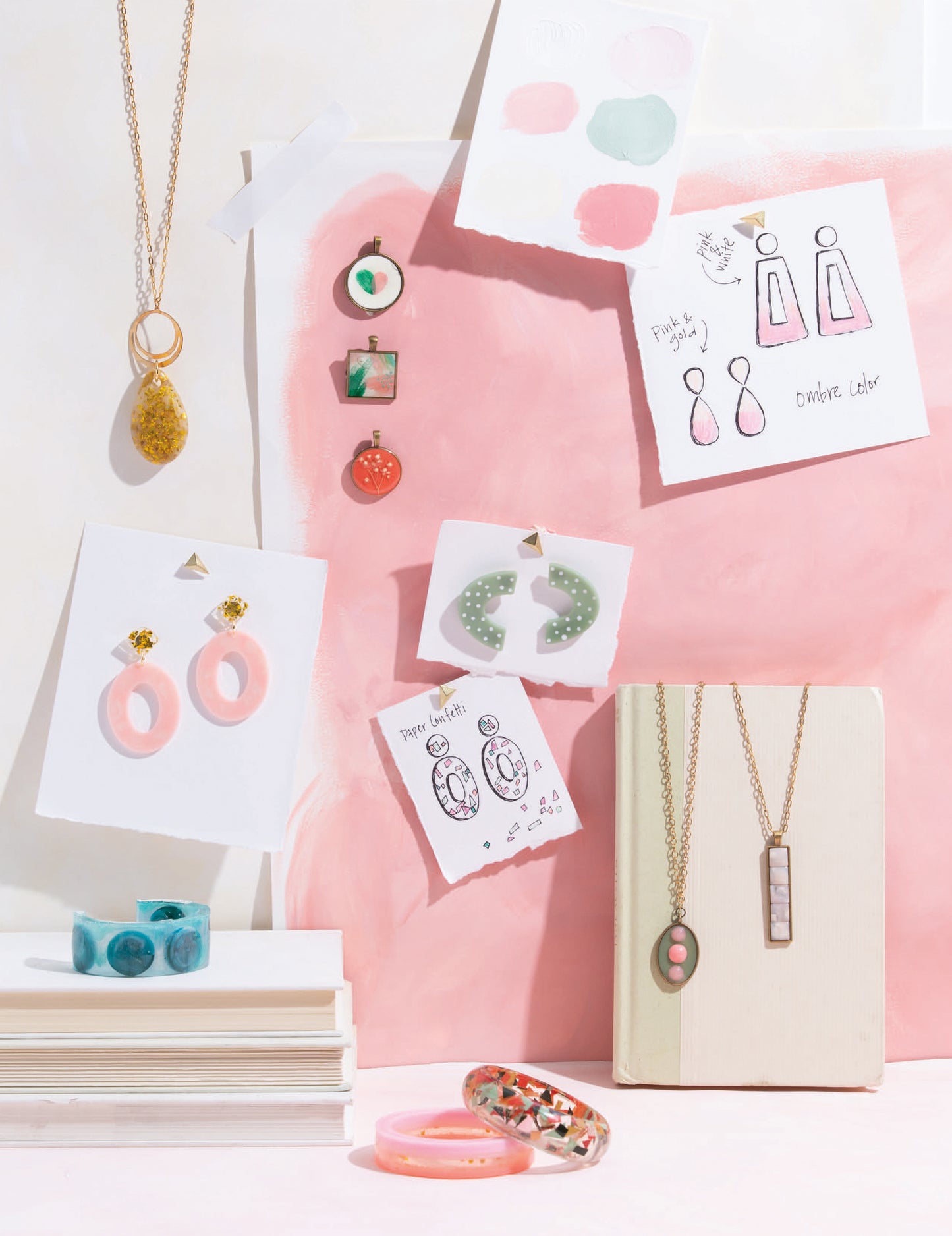 Resin jewelry, including earrings, necklaces, and rings from "Resin Jewelry—15 Fashion Forward Designs to Accessorize Your Looks" by Leisure Arts, is displayed on books with sketches and pastel swatches for UV resin accessory inspiration.