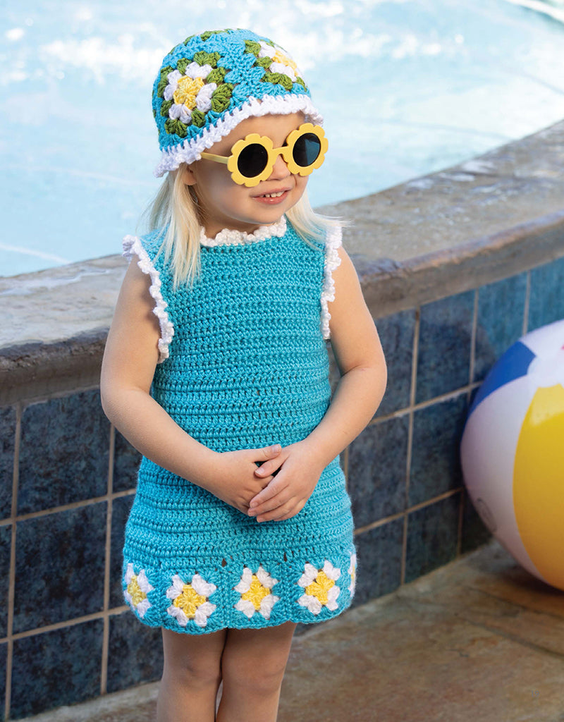 A young girl stands by a pool wearing the Great Grannies Made Modern crochet blue dress with white and yellow flower squares, matching hat, and yellow flower sunglasses from Leisure Arts. A beach ball lies on the ground beside her.