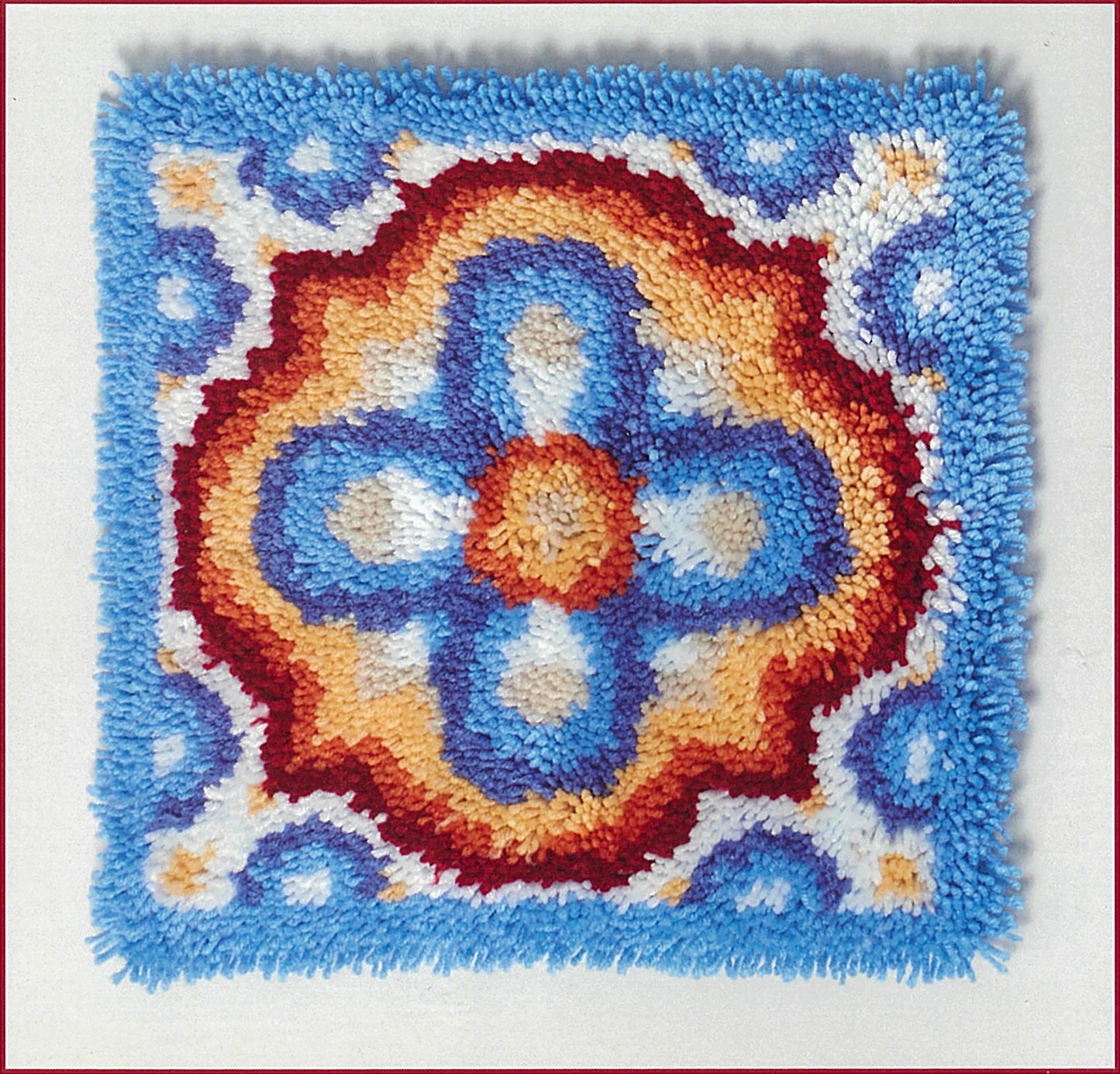 The Timeless Tiles in Latch Hook by Leisure Arts features a square rug with a floral, clover-like pattern in blue, orange, red, white, and yellow plus a blue border—ideal for beginners exploring new latch hook designs.