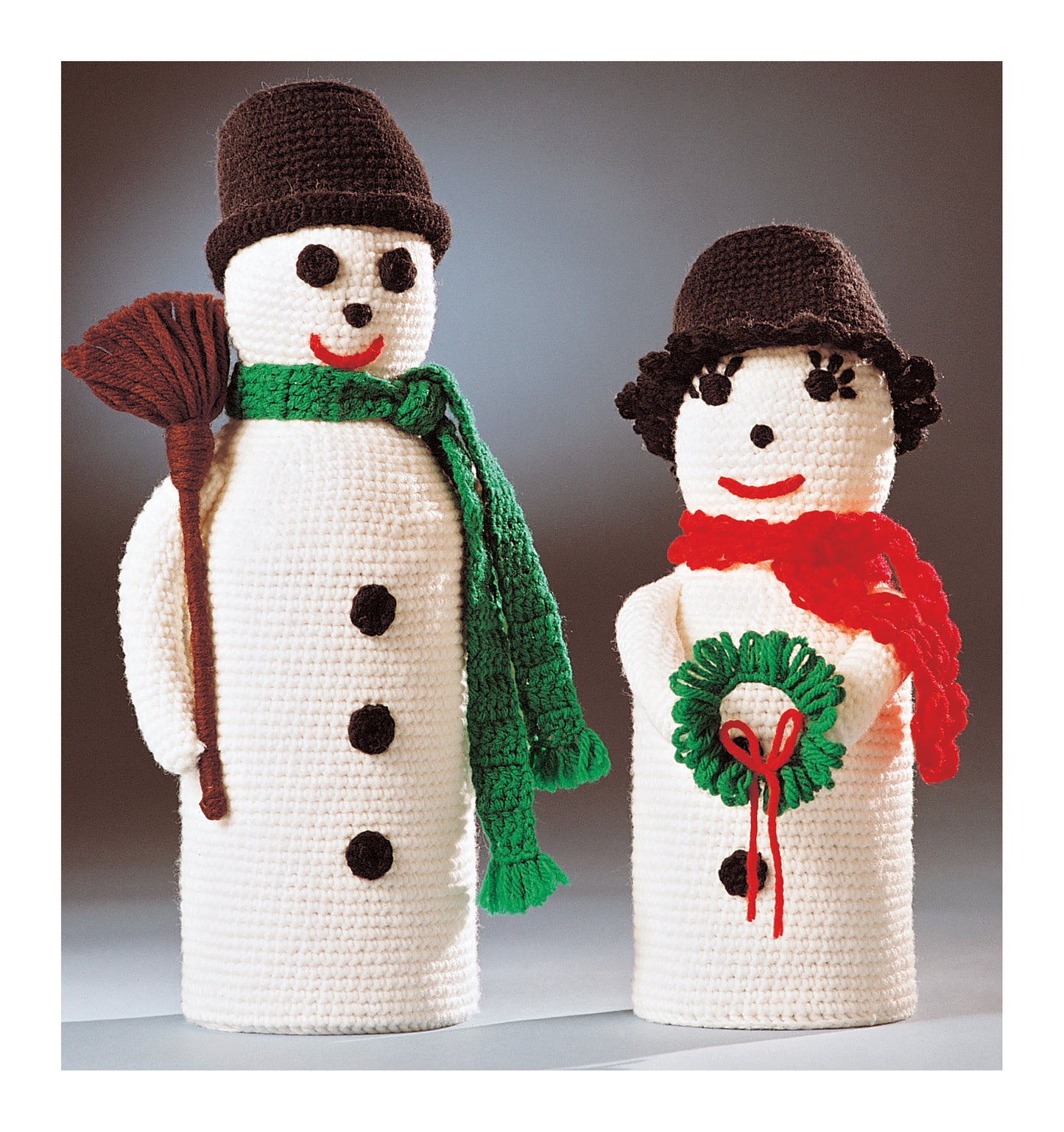 Crochet for Christmas by Leisure Arts features two smiling crocheted snowmen in black hats; one holds a broom with a green scarf, the other wears a red scarf and carries a green wreath—charming home decor craft accents.