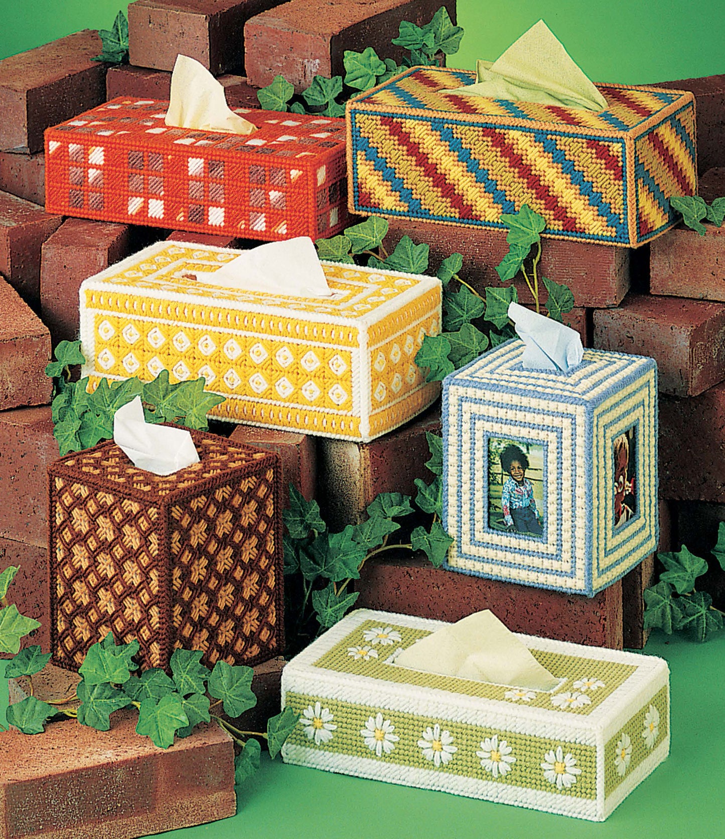 Six vibrant Leisure Arts Tissue Box Covers for Plastic Canvas are hand-stitched with intricate patterns, displayed among red bricks and green ivy on a green background.