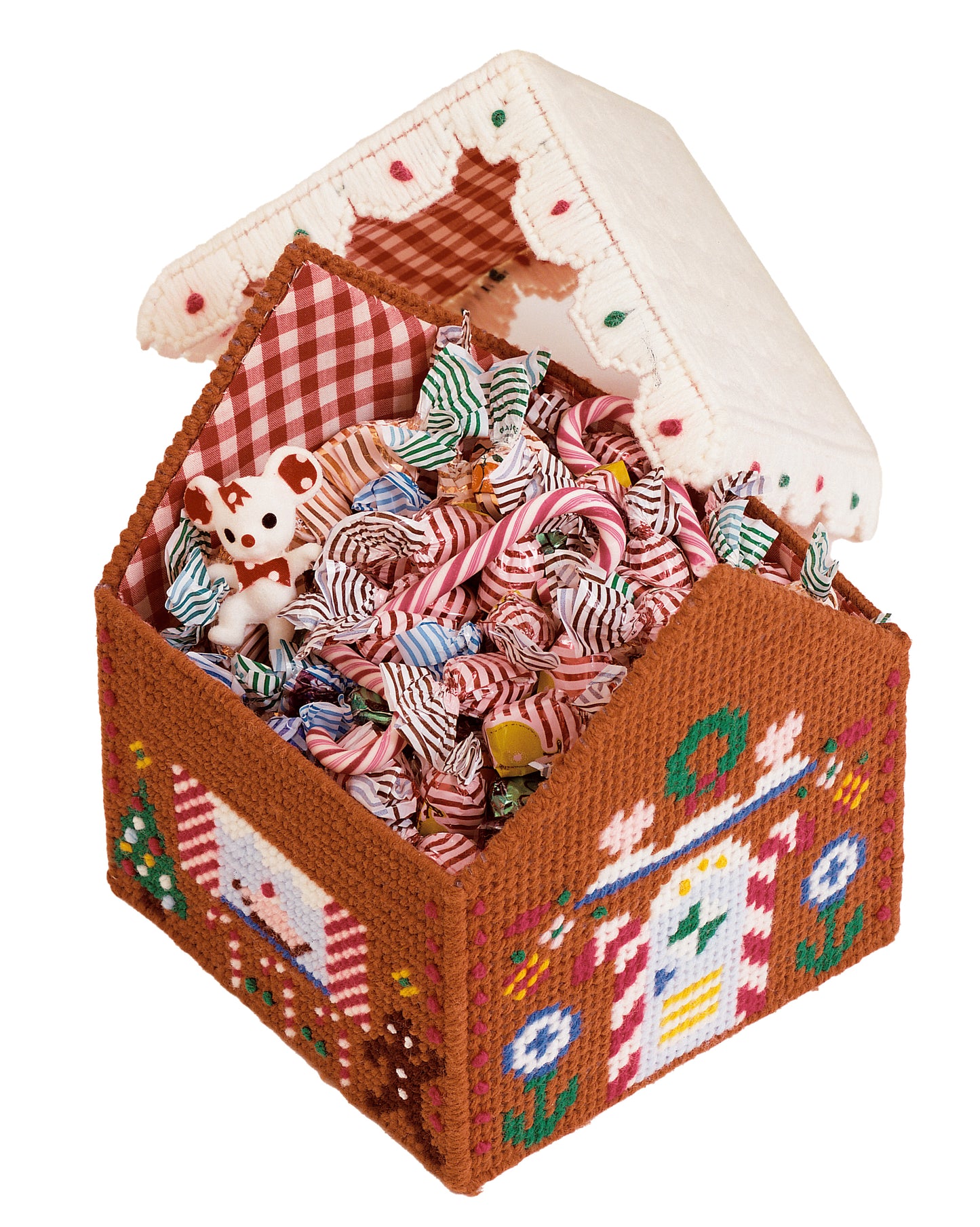 The Leisure Arts "More Christmas Projects for Plastic Canvas" features a hand-stitched gingerbread house box, ideal for beginners, shown open with festive red-and-white candies, a stuffed bear, and gingham cloth inside.