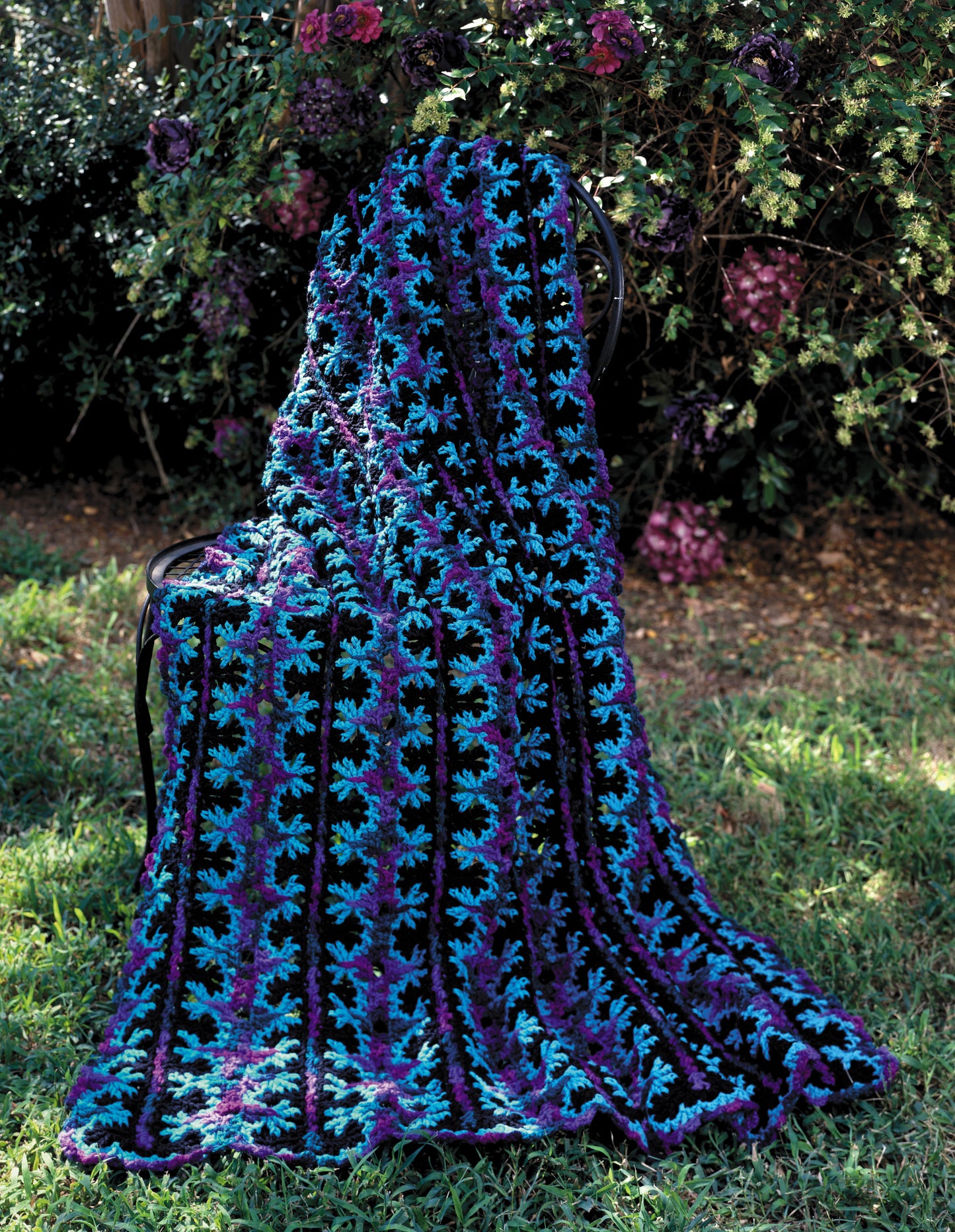 The Contest Favorite Mile-A-Minute Afghans Digital Download by Leisure Arts features a vibrant crocheted blanket in blue, teal, purple, and black, draped over a garden chair amid green grass and blooming purple flowers.