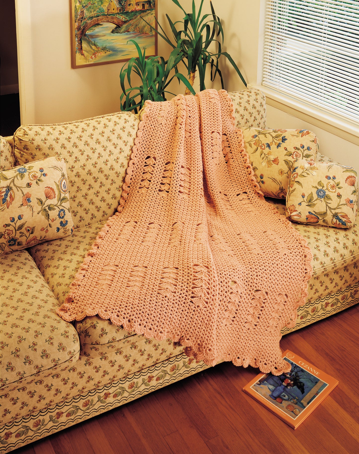 The “Afghan Parade” by Leisure Arts, crafted in soft salmon pink worsted yarn, is draped over a floral beige couch with matching pillows. A magazine rests on the wood floor; a houseplant and framed painting complete the cozy scene.