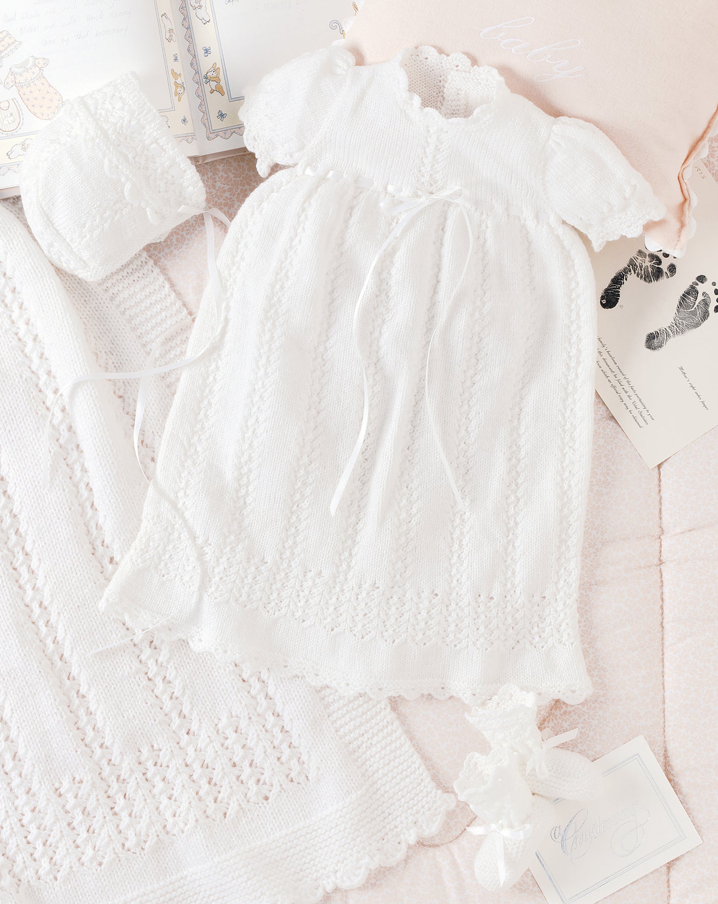 The Leisure Arts Knit Layette, featuring a delicate white knitted baby dress with short sleeves and ribbon ties, is displayed with a matching bonnet, blanket, and card among open baby books and a pink pillow on a pastel background.
