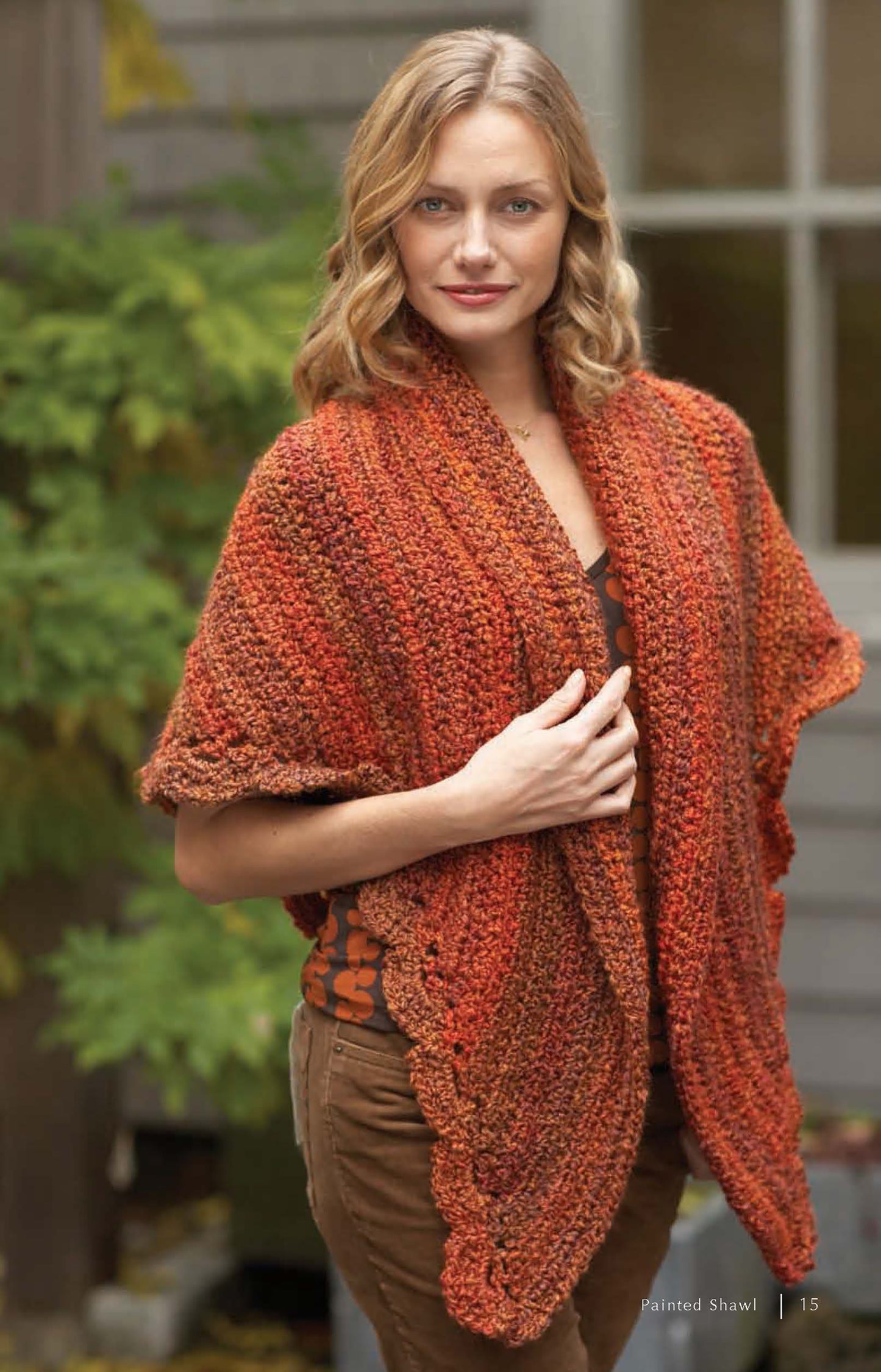 A woman with wavy blonde hair stands outdoors in front of greenery, wearing a Cozy Cover-Ups shawl from Leisure Arts. The design features autumn shades and is made with Lion Brand Homespun yarn. Digital download includes 6 crochet and knit patterns.