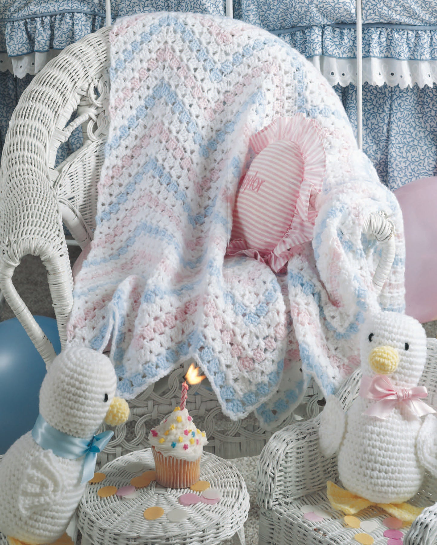 Just Ducky Baby Afghans by Leisure Arts drape over a white wicker chair, surrounded by crochet ducks, pastel balloons, and a cupcake with a lit candle—perfect for charming nursery gifts.