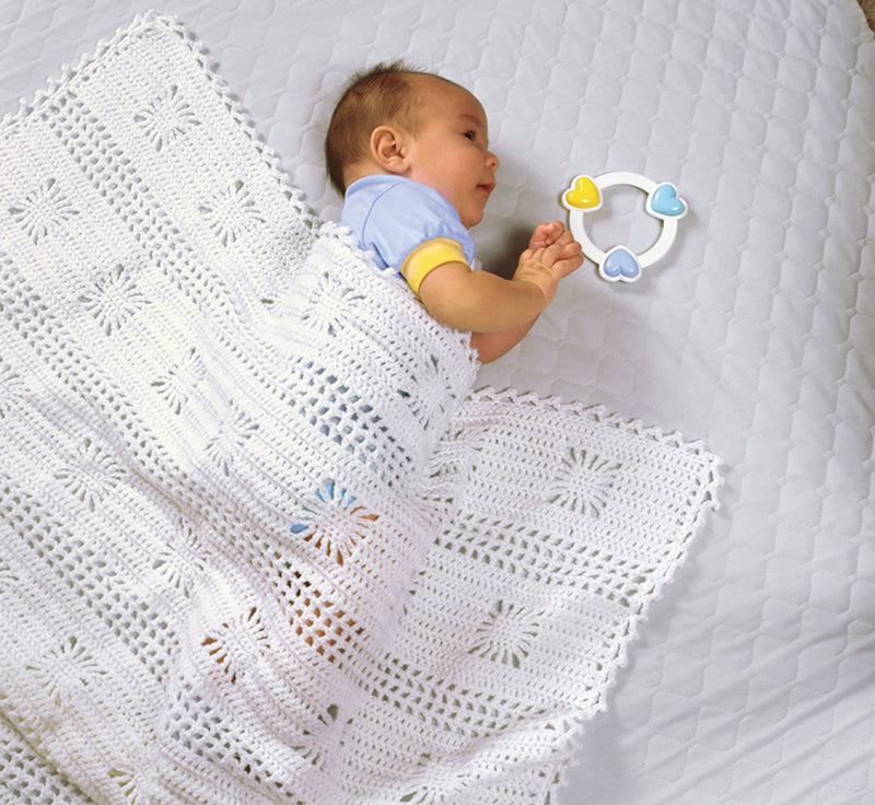 A baby rests on a white quilt, snuggled under a Make One for Baby crochet blanket by Leisure Arts, gazing at a circular rattle with heart-shaped ends in blue and yellow—perfect inspiration for beginner crochet patterns.