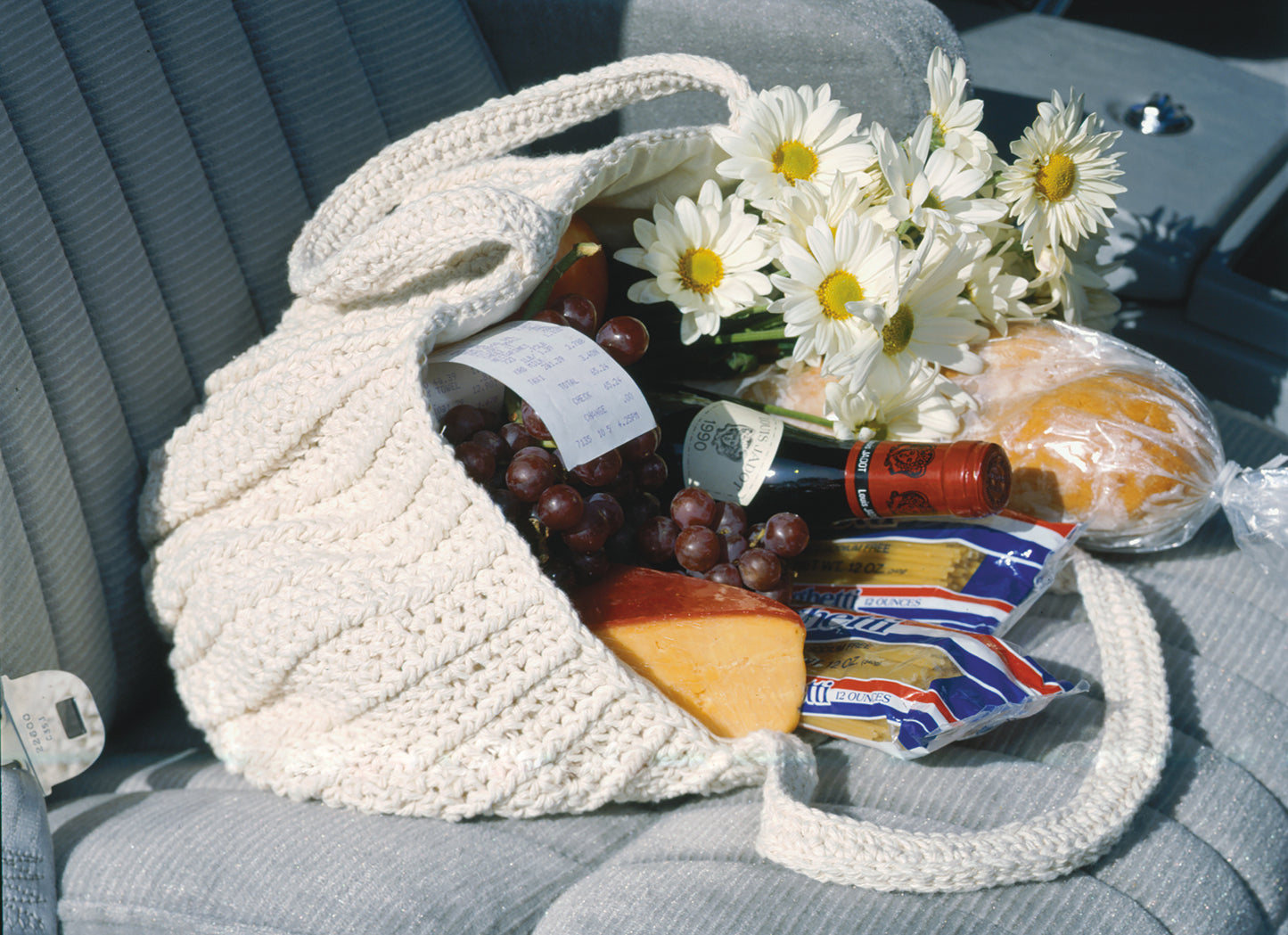 The Enviro-Totes Digital Download by Leisure Arts showcases a white cotton crochet bag on a car seat, filled with groceries—grapes, wine, cheese, pasta, bread, daisies—while a printed receipt peeks out.