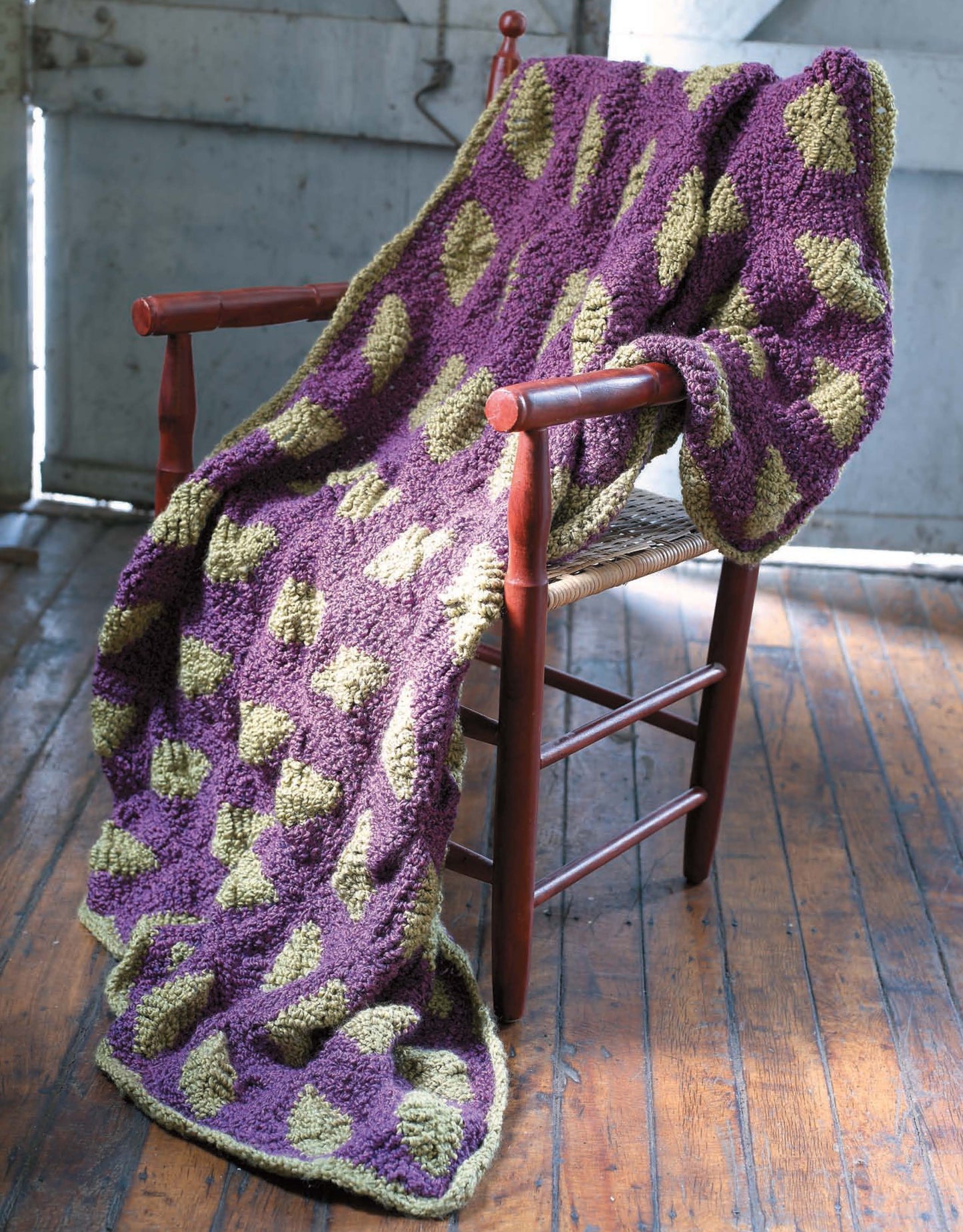 The Story of Homespun Crochet by Leisure Arts features a purple and green bulky knit blanket, elegantly draped over a wooden chair on a sunlit floor with a rustic, weathered wall as the backdrop.