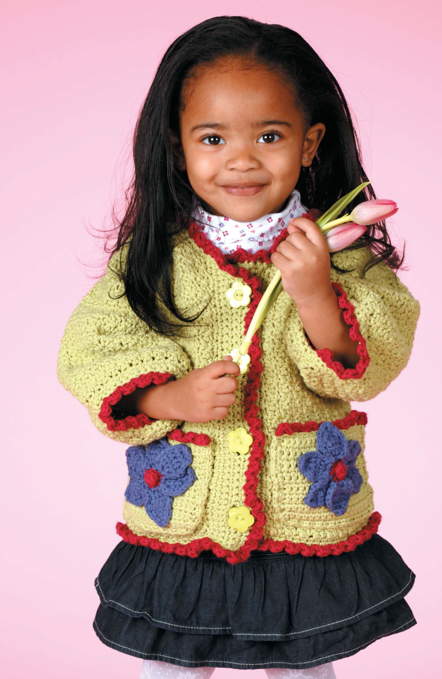A young girl in Leisure Arts’ Crochet Sweaters & Dresses for Girls—a yellow floral sweater and black skirt—smiles holding two pink tulips against a pink background.