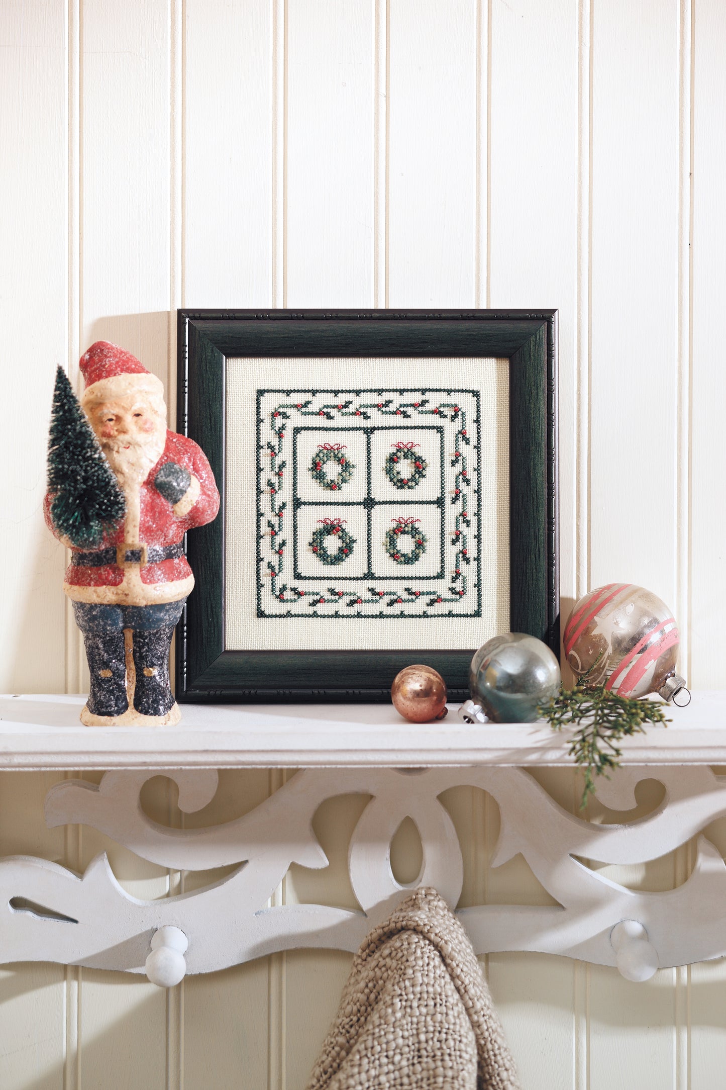 A shelf holds the Leisure Arts "Sweet Nothings Cross Stitch for Christmas," a vintage Santa with tree, Christmas ornaments, and greenery, all set against a white paneled wall.