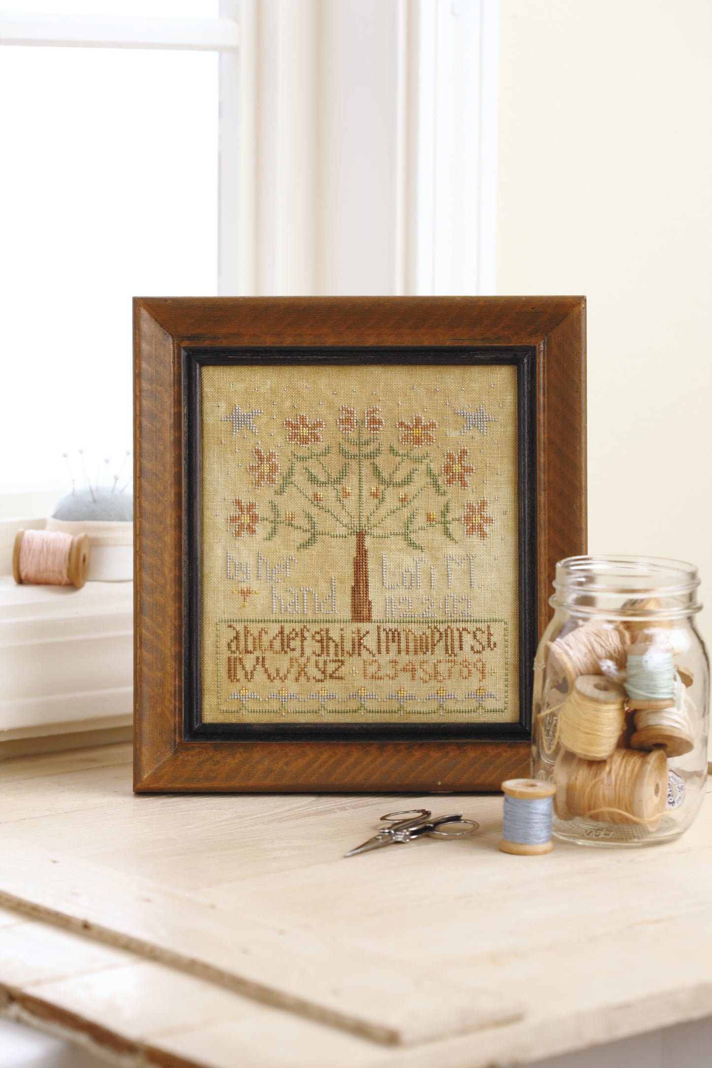 The "Words To Stitch By" cross-stitch sampler by Leisure Arts features an alphabet, numbers, and floral tree, displayed on a table with sewing tools—ideal for anyone who loves beautiful needlework and creative patterns.