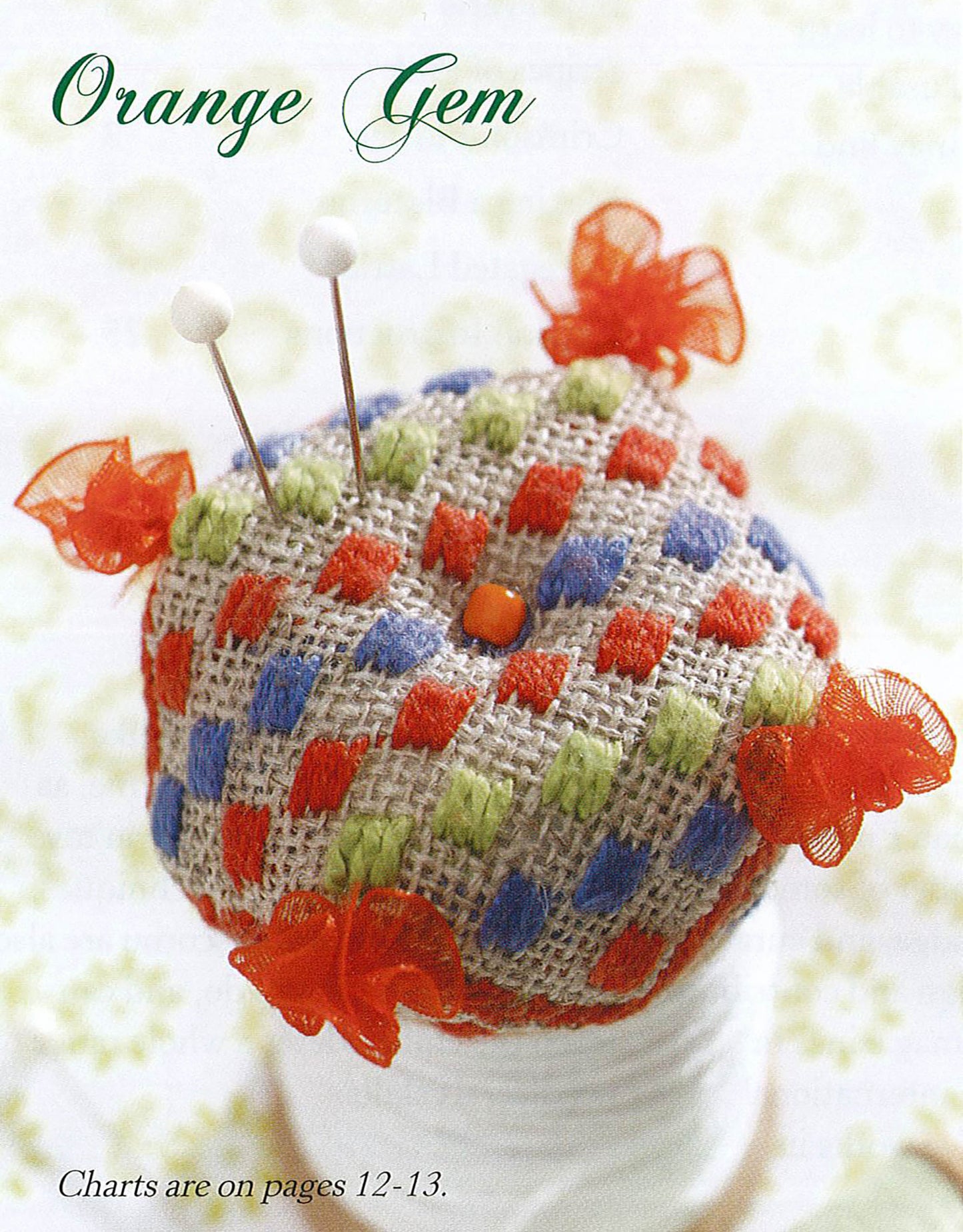 The "Teach Yourself to Make Biscornu Digital Download" by Leisure Arts features round, woven pincushions with orange, green, and white checkered patterns—ideal for cross stitch or embroidery. Shown atop a spool of white thread.