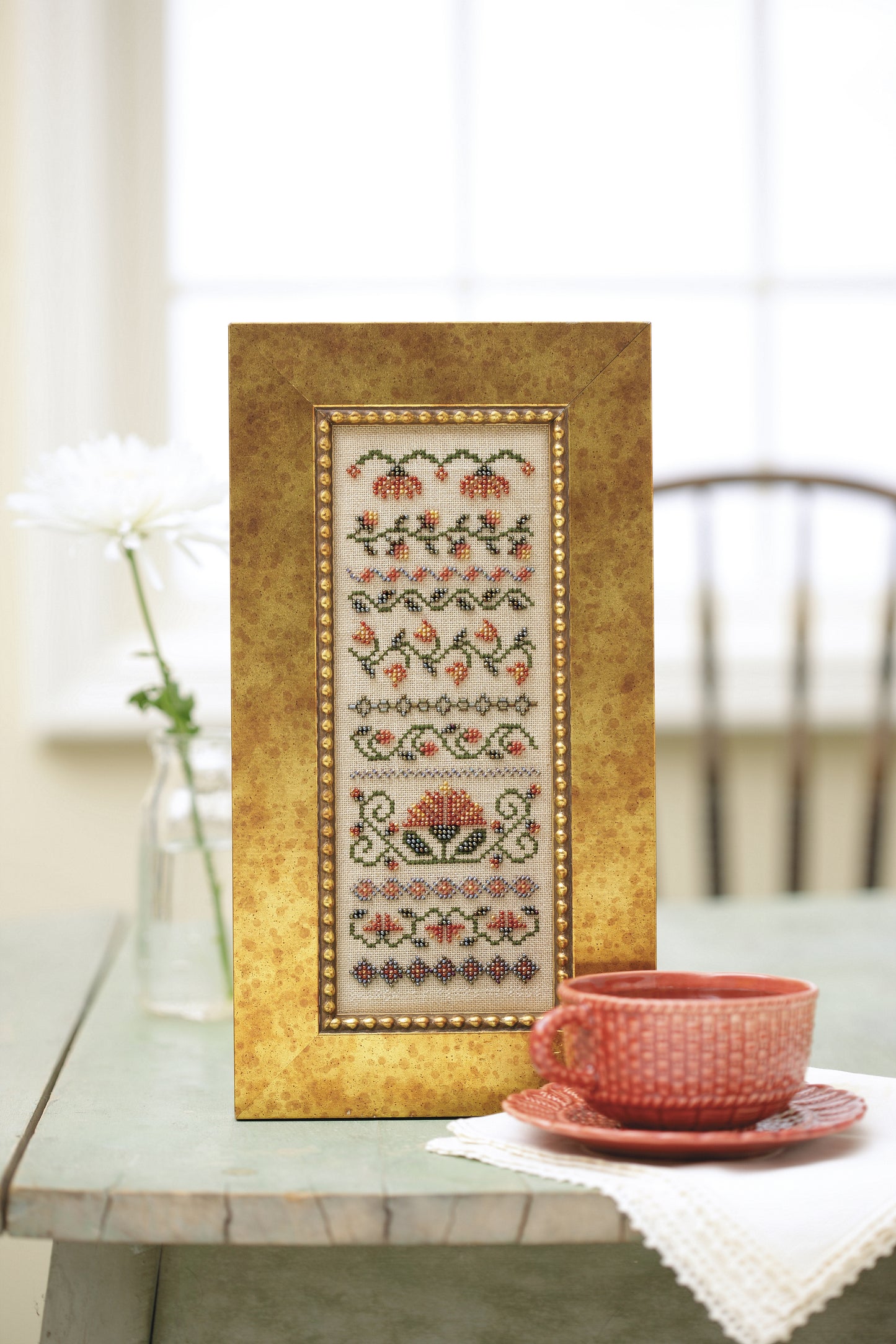 A framed beaded floral embroidery from Stitch a Beaded Garden—18 Designs Charted for Beading and Cross Stitch (Leisure Arts) is displayed by a pink teacup, saucer, and white flower in a vase, with a chair and sunny window behind.