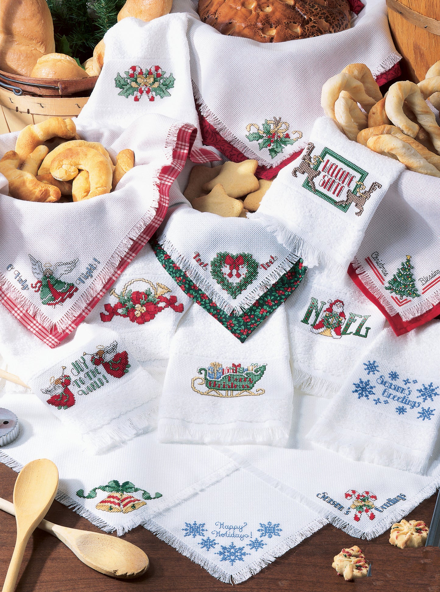 A festive display of Leisure Arts' Christmas Towels & Bread Cloths, featuring embroidered holiday patterns like wreaths and snowflakes, arranged with breads, cookies, and wooden utensils for a charming seasonal touch.