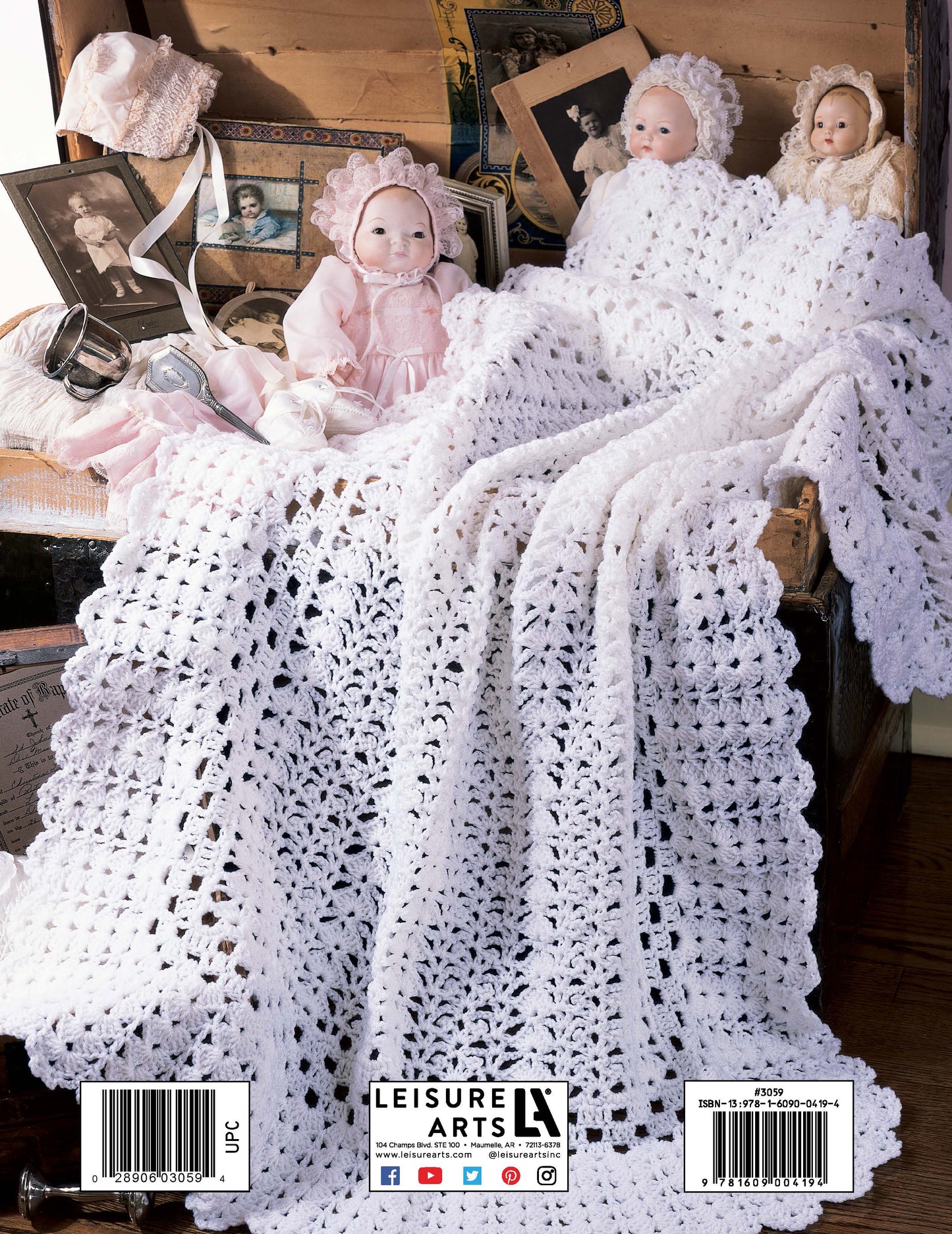 A white crocheted blanket from Leisure Arts' Heirloom Afghans for Baby drapes over an open chest filled with vintage dolls and photos, creating a nostalgic, heirloom-inspired scene.