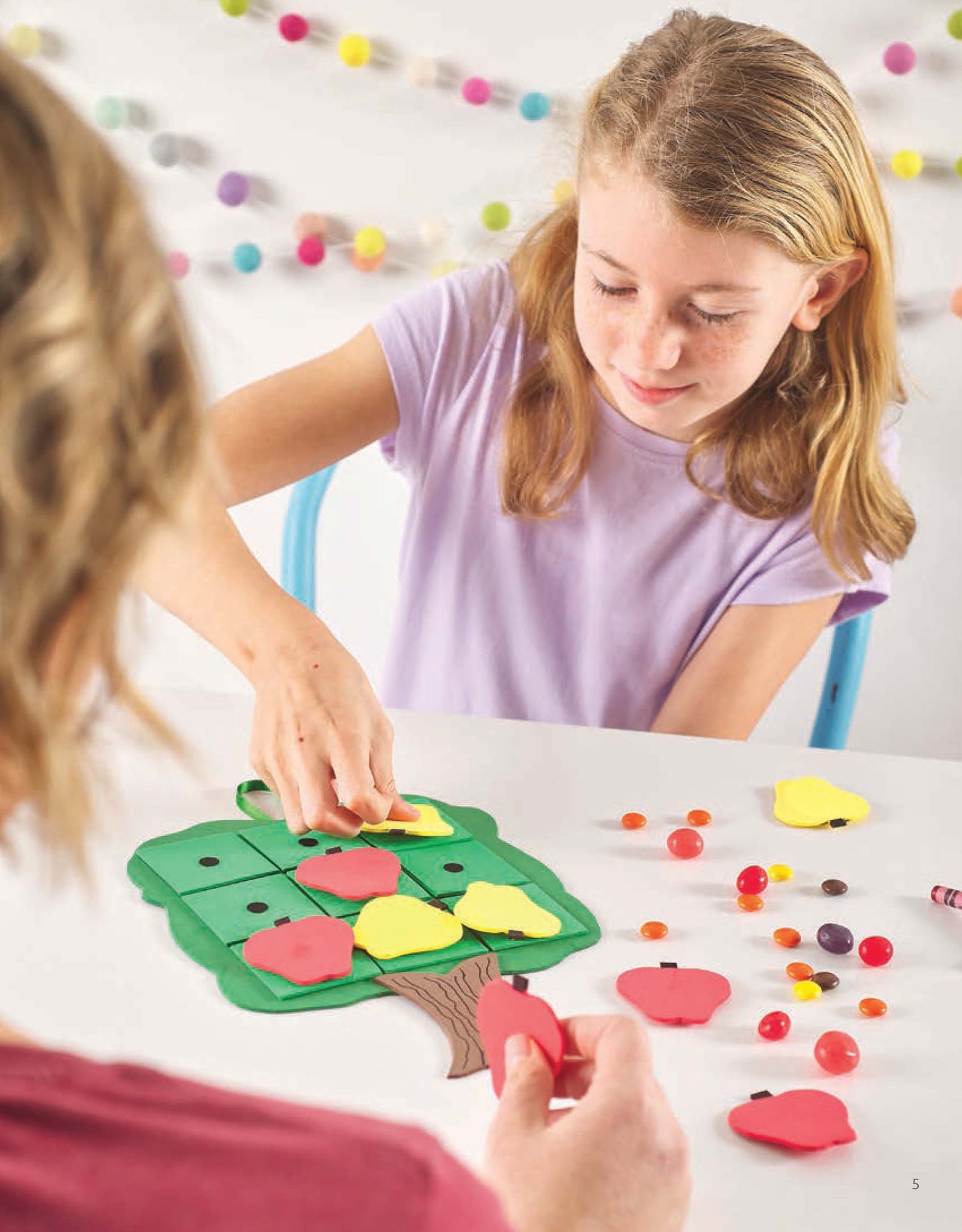 Two children play a tabletop game, placing apples on a felt tree amid colorful beads and garlands. One child enjoys creative hands-on fun with Craft Foam Favorites by Leisure Arts, perfect for inspiring kids’ crafts.