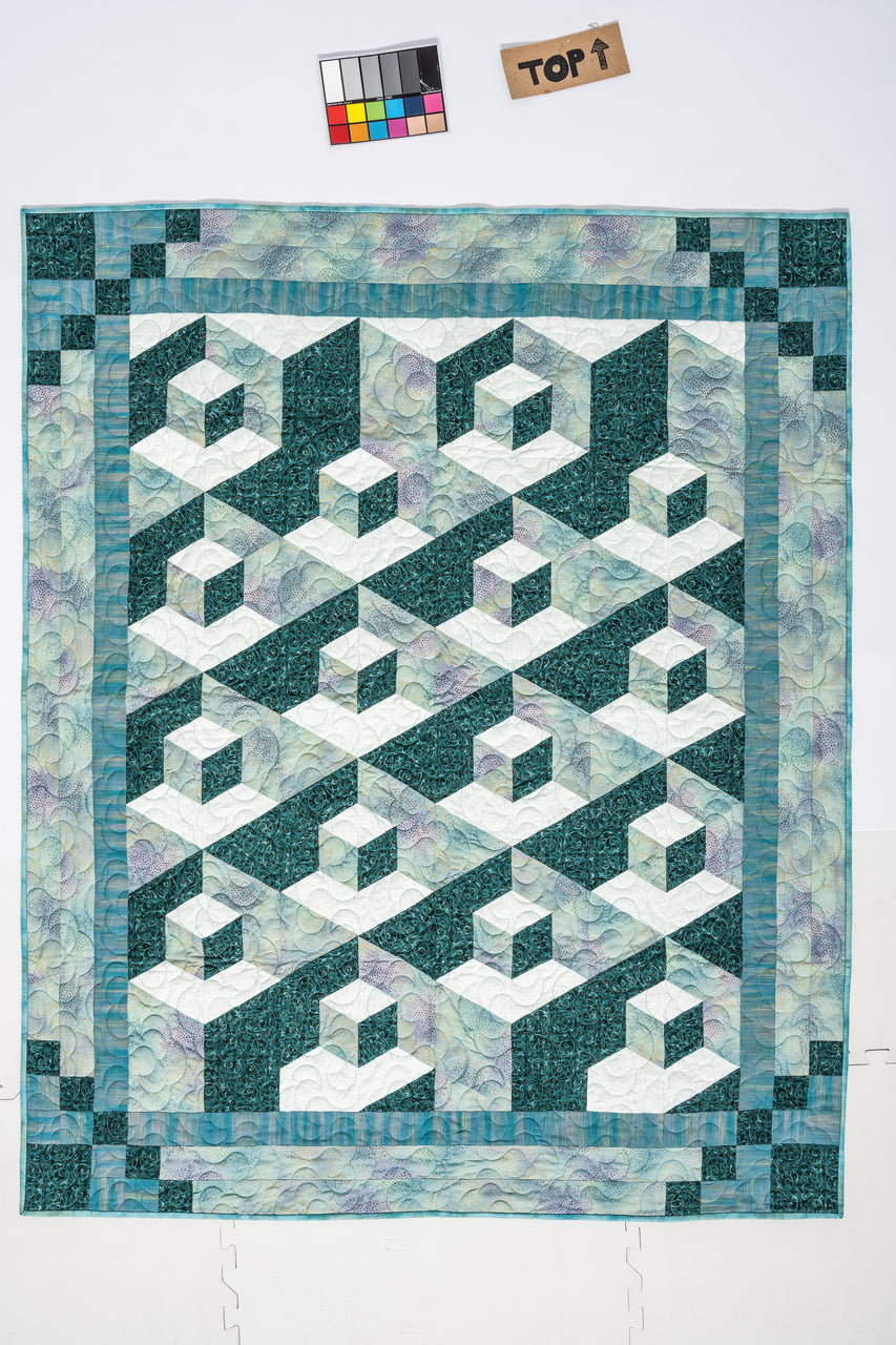 The quilt from C&T Publishing’s 3-D Magic! Simple Blocks, Striking Quilts Digital Download features a geometric 3D cube pattern in teal, white, and aqua with a teal border and pastel swirled background. A color checker and TOP sign rest above.