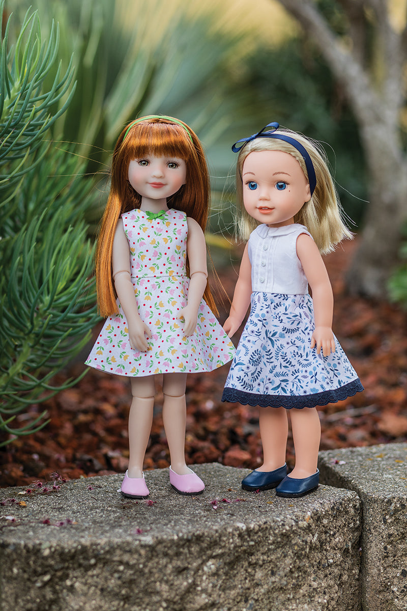 Two dolls stand on stone outdoors, modeling 14" outfits made with Doll Studio Boutique Digital Download sewing patterns by FunStitch Studio—one in a pastel dress, one in blue and white—against green plants.