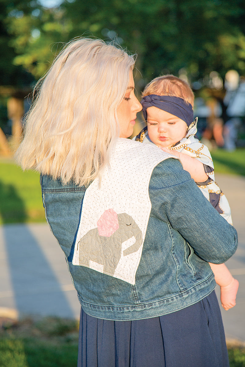 A woman outdoors holds a baby and displays a white cloth featuring Sew Cute Baby Animals Digital Download by C&T Publishing—ideal for nursery decor or baby gifts. The scene includes trees, a sidewalk, and both wear adorable outfits.