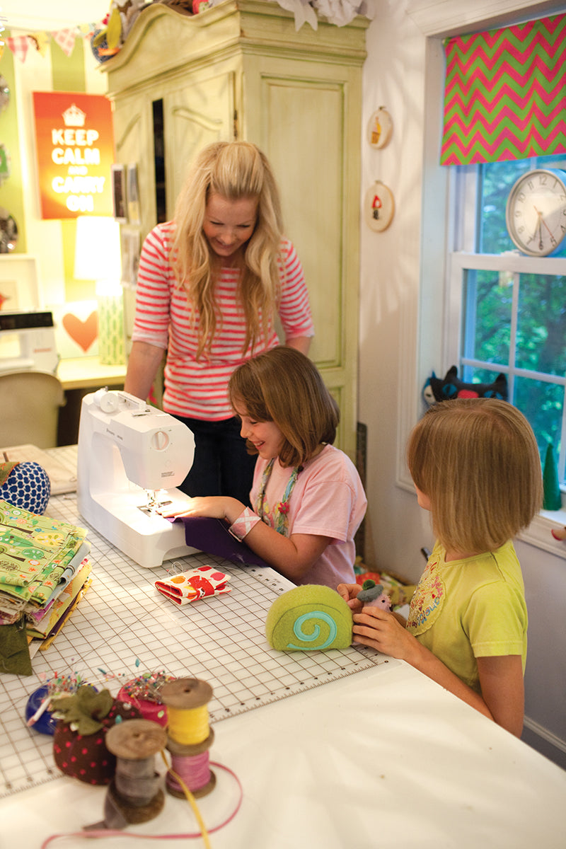 A woman and two girls use a sewing machine together at a colorful table, learning with the Sewing for Beginners Handy Pocket Guide Digital Download from C&T Publishing. The bright room is filled with fabric and supplies.