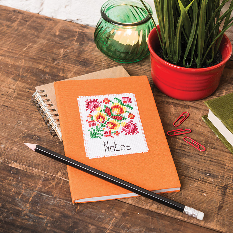A modern folk-art cross stitch pattern from C&T Publishing’s "Modern Folk-Art Cross Stitch Digital Download," labeled Notes, is displayed on an orange notebook placed on a wooden desk with office supplies and a potted plant.