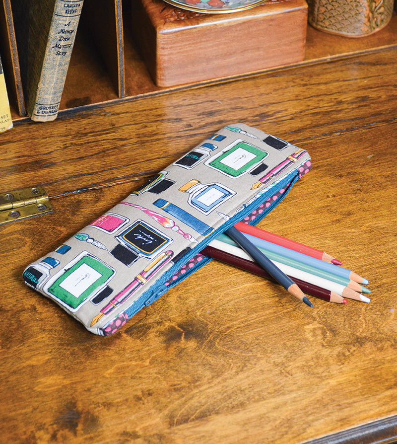 A fabric pencil case with a book-themed pattern sits open on a wooden desk, colored pencils spilling out—a charming scene inspired by the Jump Into Sewing Digital Download from C&T Publishing. Books and decor add color to the background.