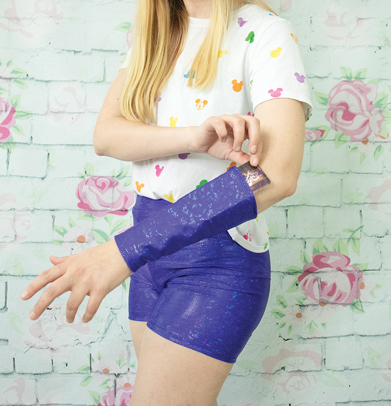 A person in a white shirt with colorful Mickey Mouse shapes and shiny purple shorts adjusts a matching arm sleeve—perfect costume inspo. Discover more with the Cosplay Foundations Digital Download from FanPowered Press! Pink floral wall in the background.