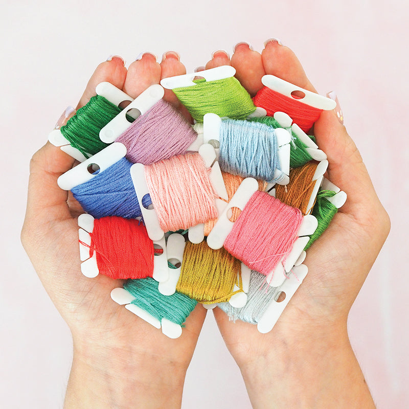 A pair of hands holds colorful embroidery floss bobbins, perfect for cross stitch, shown with Jump Into Cross Stitch Digital Download by C&T Publishing against a soft pink background.