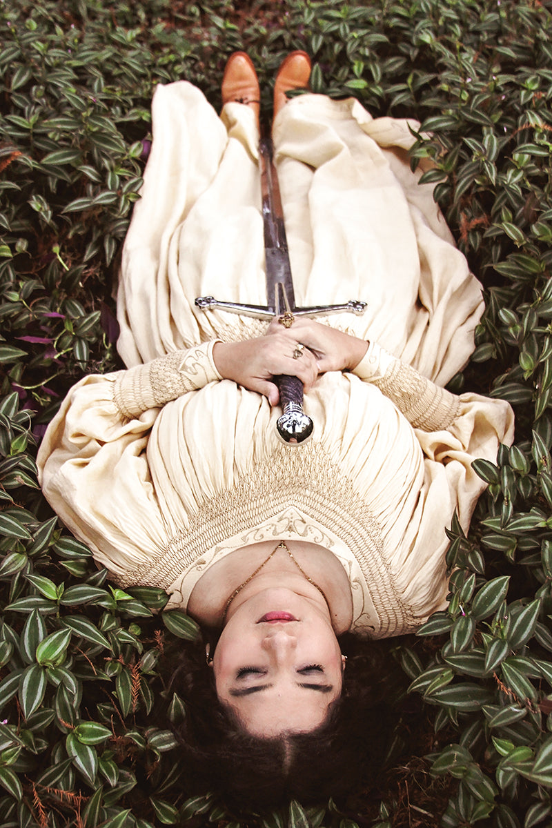 A person in a cream-colored, vintage-style dress created using FanPowered Press’s Smocking Secrets Digital Download lies on their back in green foliage, eyes closed, holding a sword with both hands on their chest, brown shoes visible.