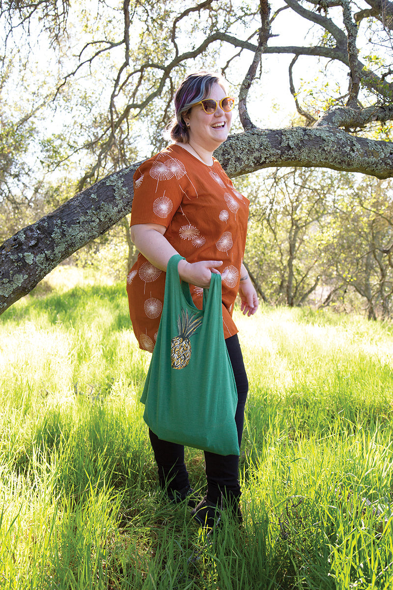 A smiling person with short hair, in sunglasses, an orange shirt, and black pants stands in a sunlit field holding a green pineapple tote—just the project you'll find in Professor Pincushion's Beginner Guide to Sewing by Stash Books. Tree branches frame the scene.