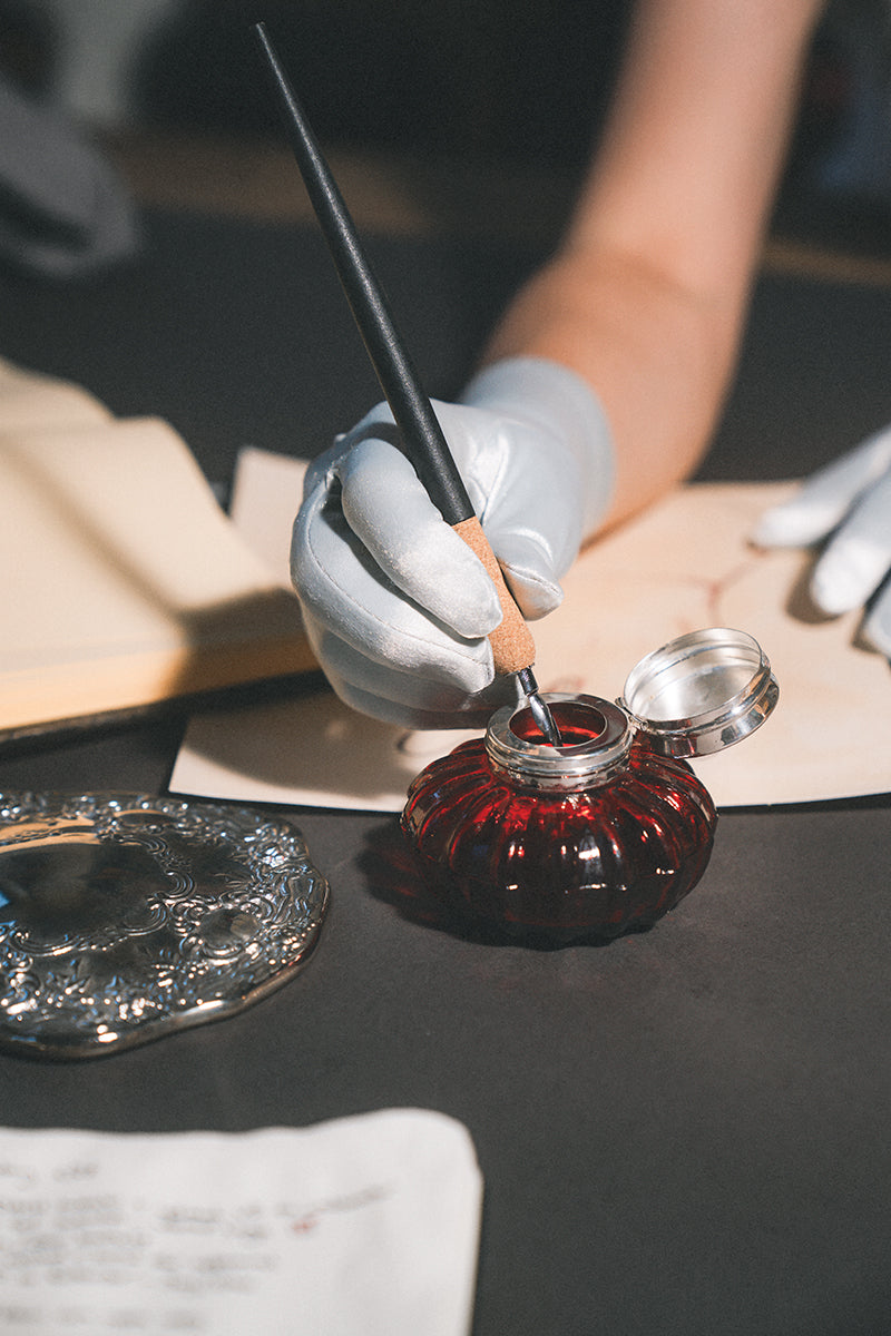 Someone explores the Handwear Handbook Digital Download by FanPowered Press, with elegant gloves, a calligraphy pen, a red glass inkwell, and an open book creating a vintage-inspired scene.